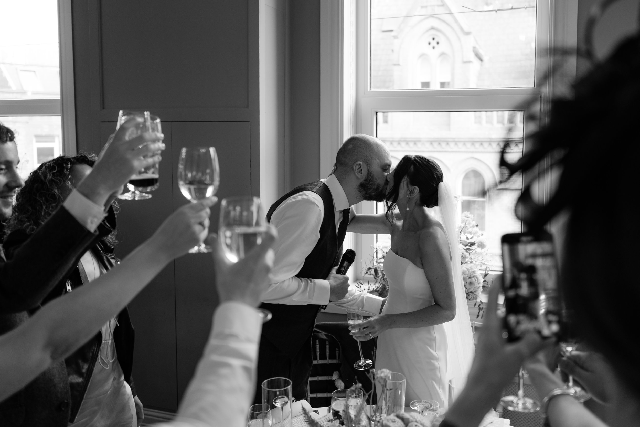 A black-and-white photo of a wedding celebration with a bride and groom kissing near a window, surrounded by guests raising glasses in a toast.