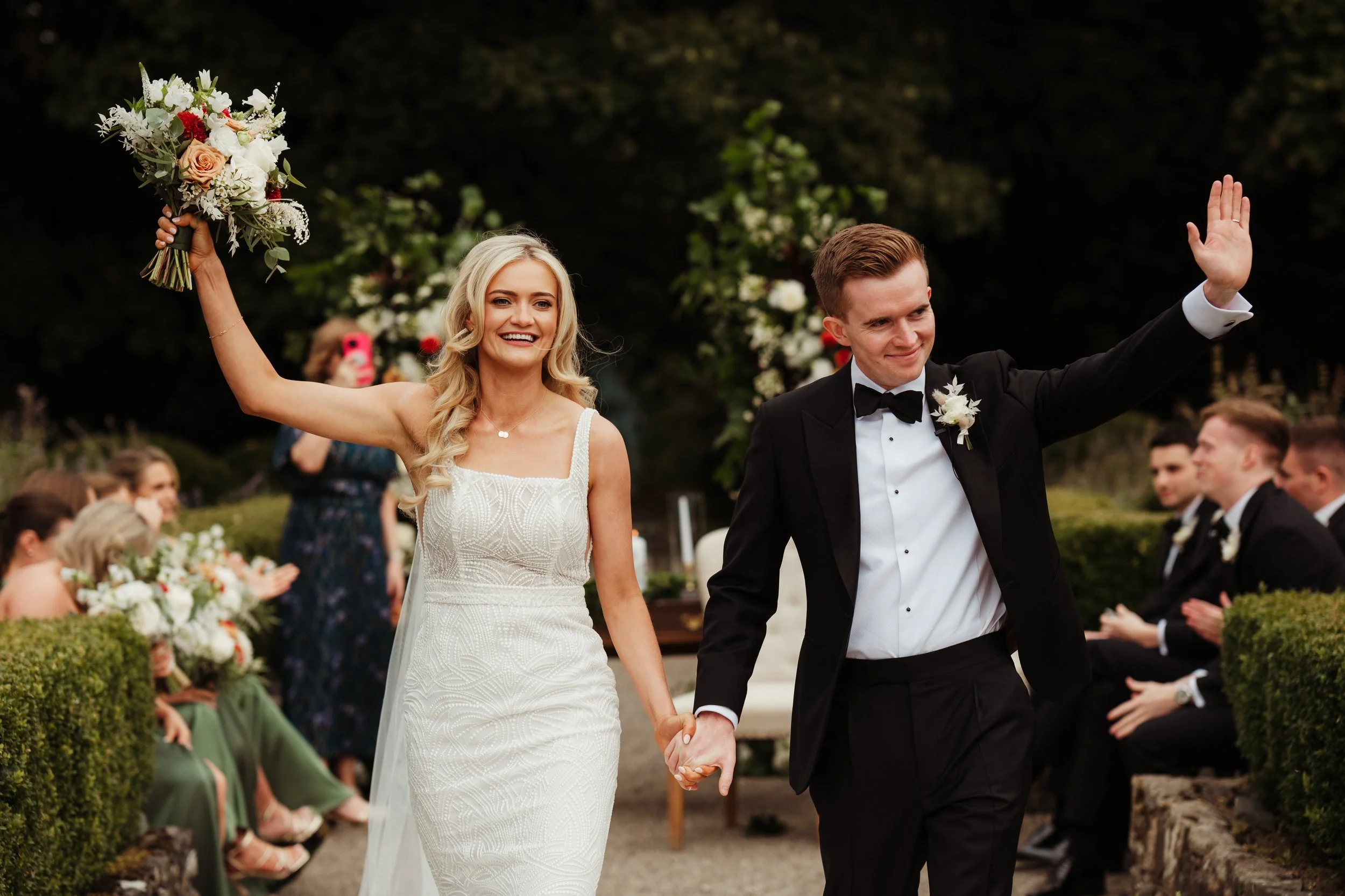 Bride holding a bouquet and smiling, groom waving, during outdoor wedding ceremony with seated guests and floral decorations in the background.