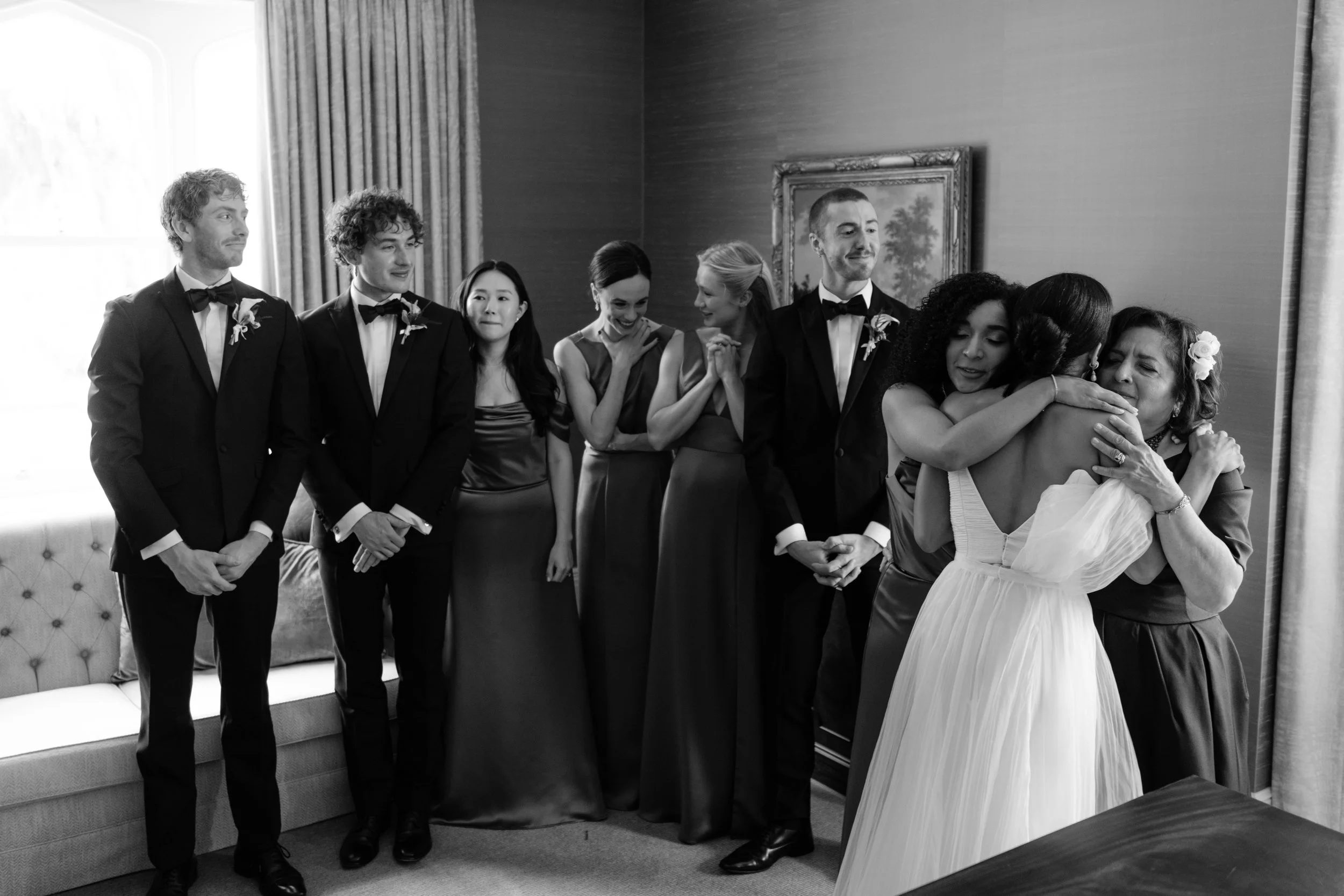 Group of people at a wedding, with two women hugging in the center, one wearing a wedding dress and the other in a dress, surrounded by friends dressed in formal attire.