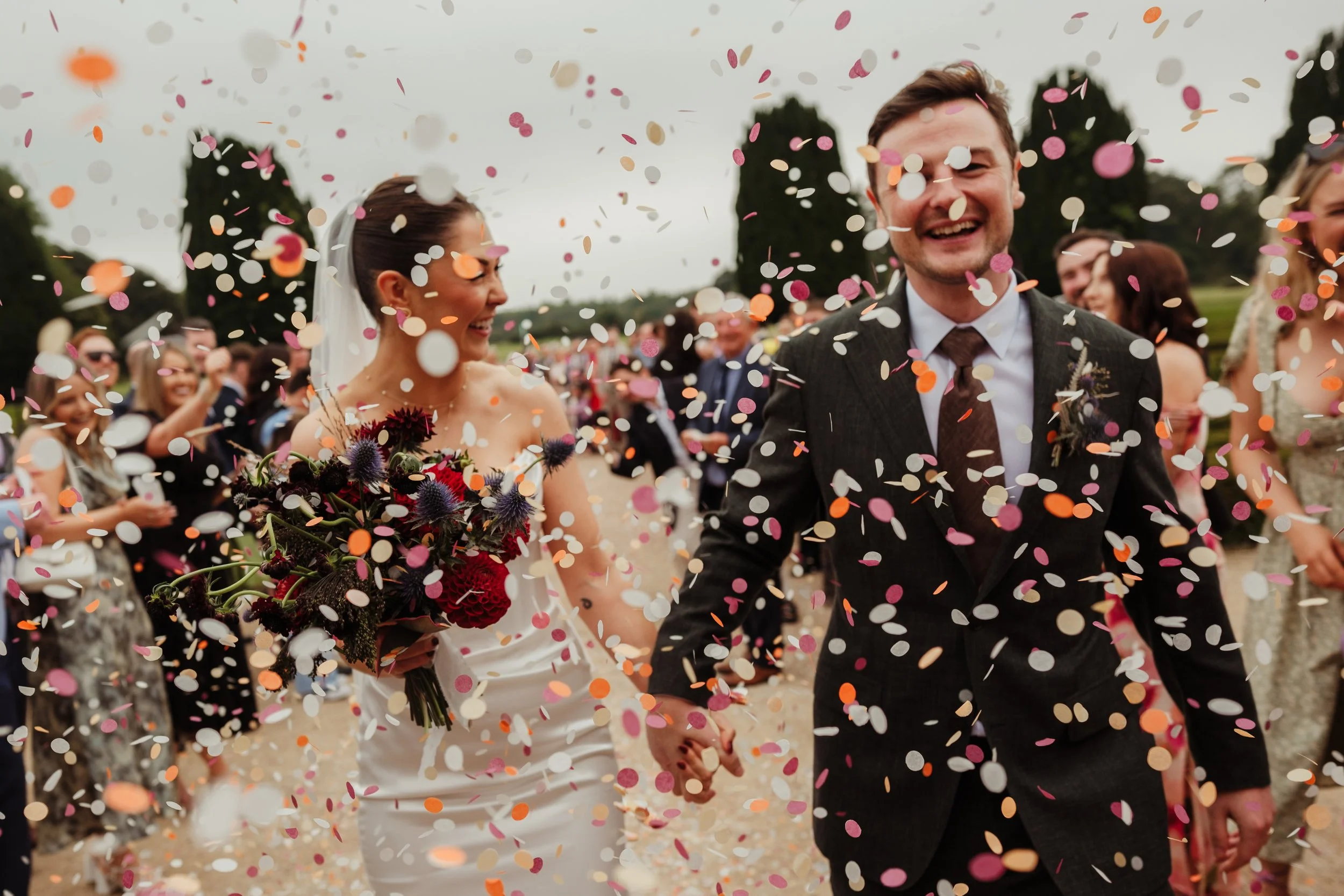 Couple in wedding attire celebrating with friends in a confetti-filled outdoor setting.