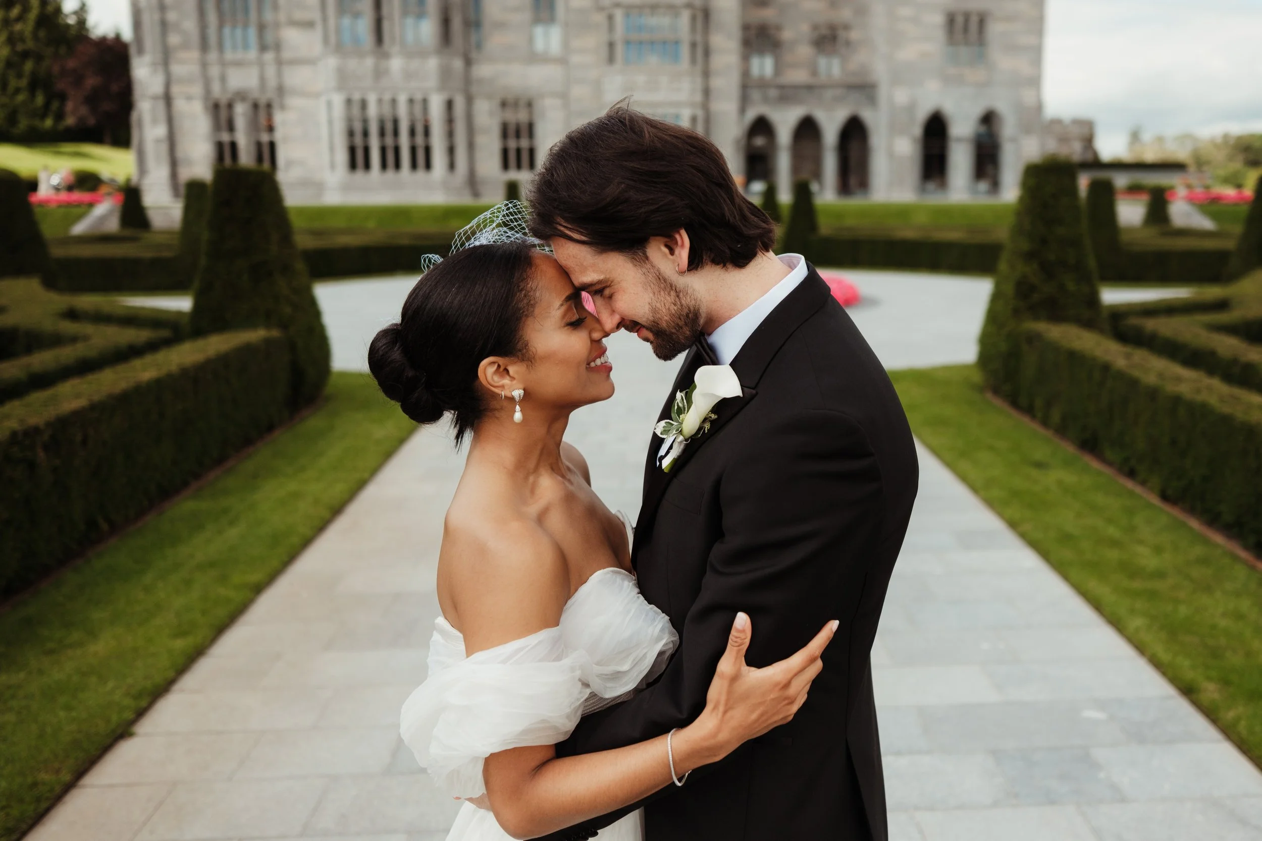A newlywed couple with foreheads touching, smiling, standing on a stone pathway in front of a historic stone building with manicured hedges and gardens.