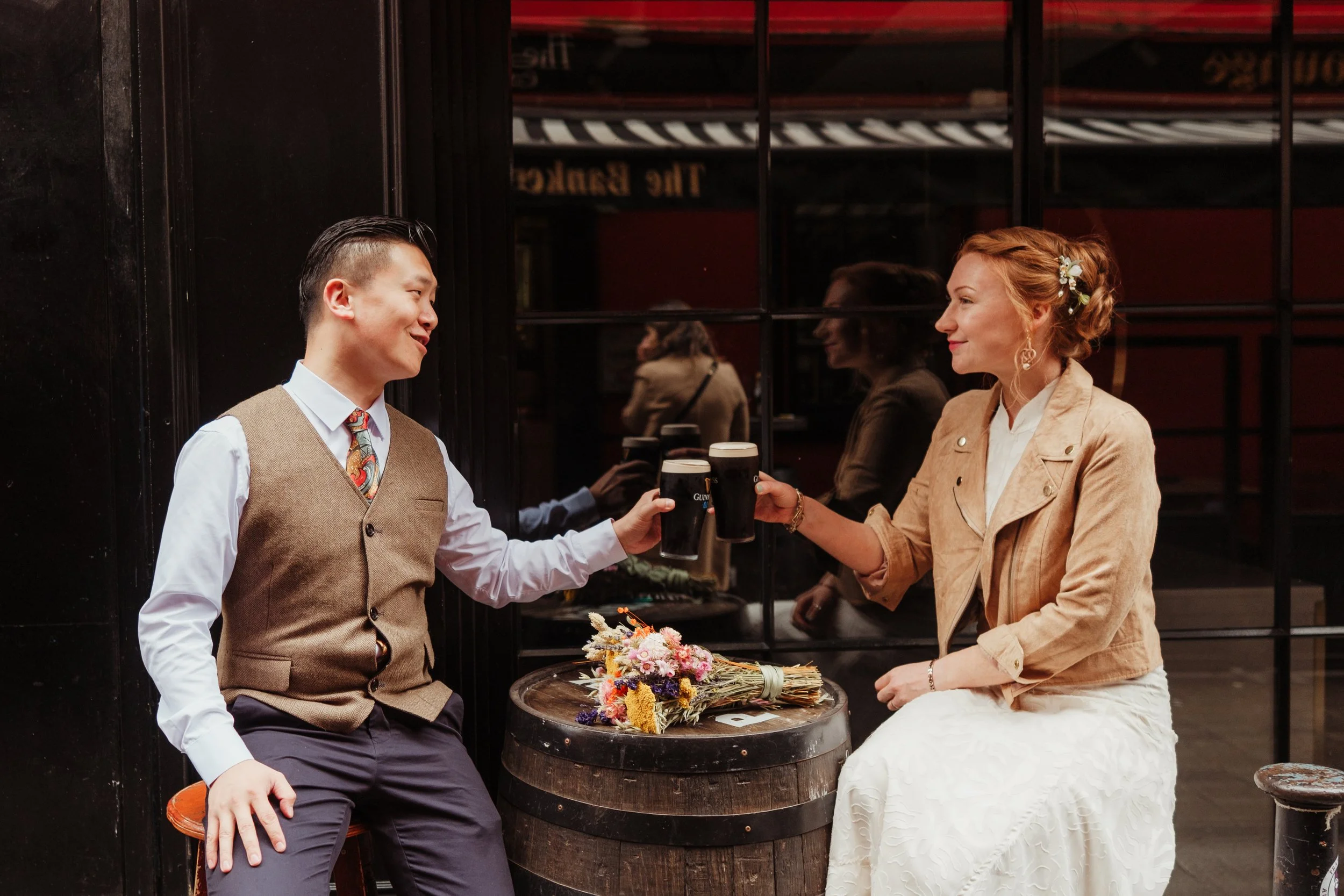 A man and woman sitting at an outdoor table, clinking pints of Guinness stout. The woman has a bouquet of flowers on the table.