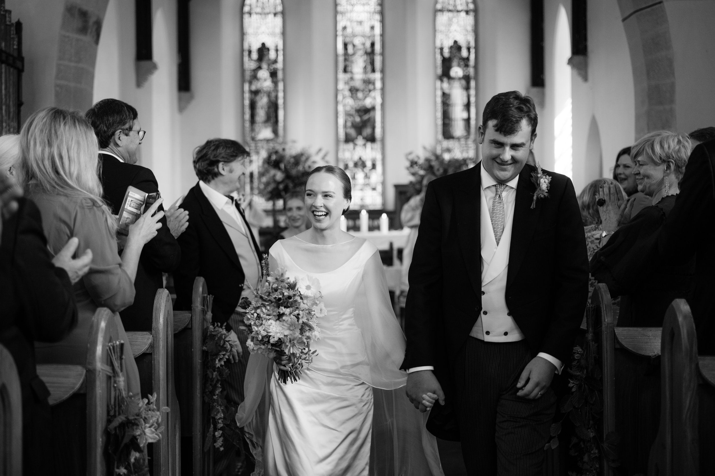 A bride and groom walking hand-in-hand down the aisle of a church, smiling and surrounded by clapping guests, during a wedding ceremony in black and white.