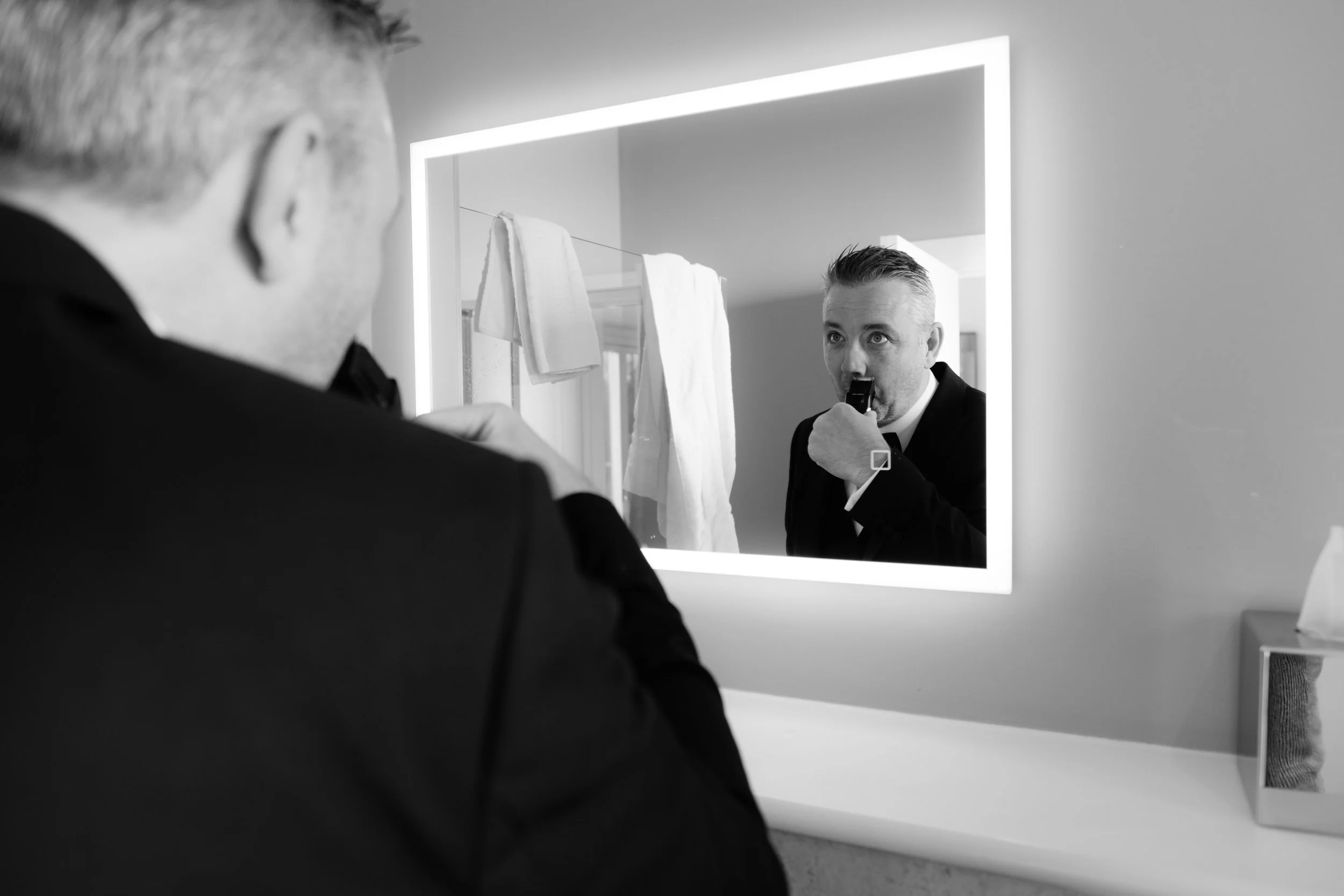 A man in a suit is looking into a mirror and holding a gun to his temple, reflected in a bathroom mirror with towel hangers and a tissue box.