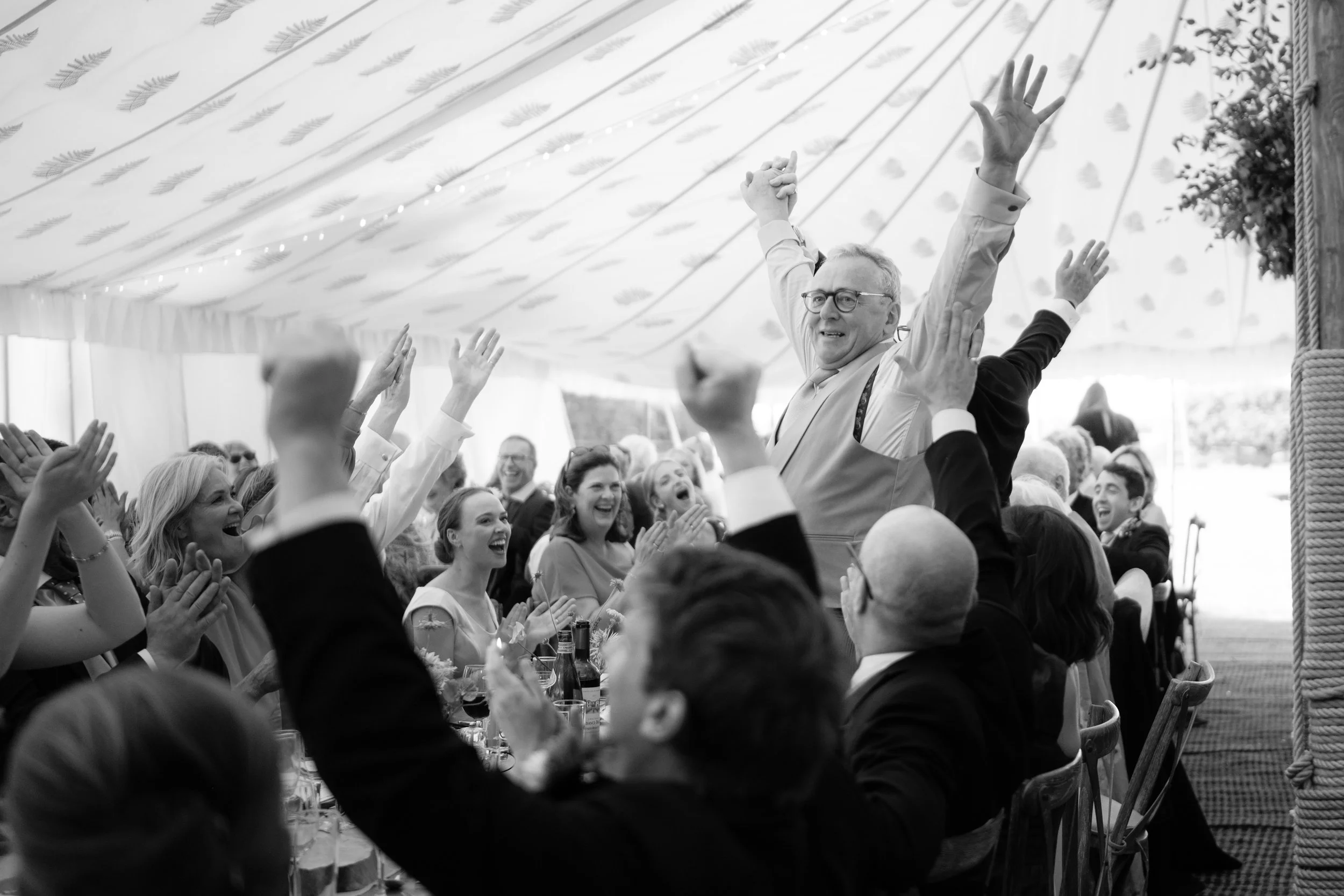 People celebrating at a festive event under a decorated tent, with some raising their hands and cheering, while an older man stands in the center raising his arms triumphantly.