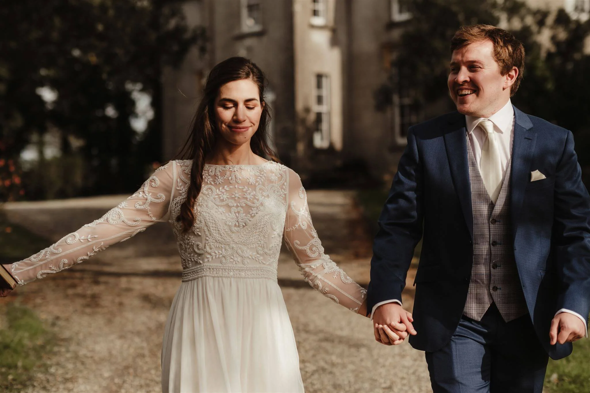 A bride and groom holding hands and smiling while walking outside in front of a historic building.