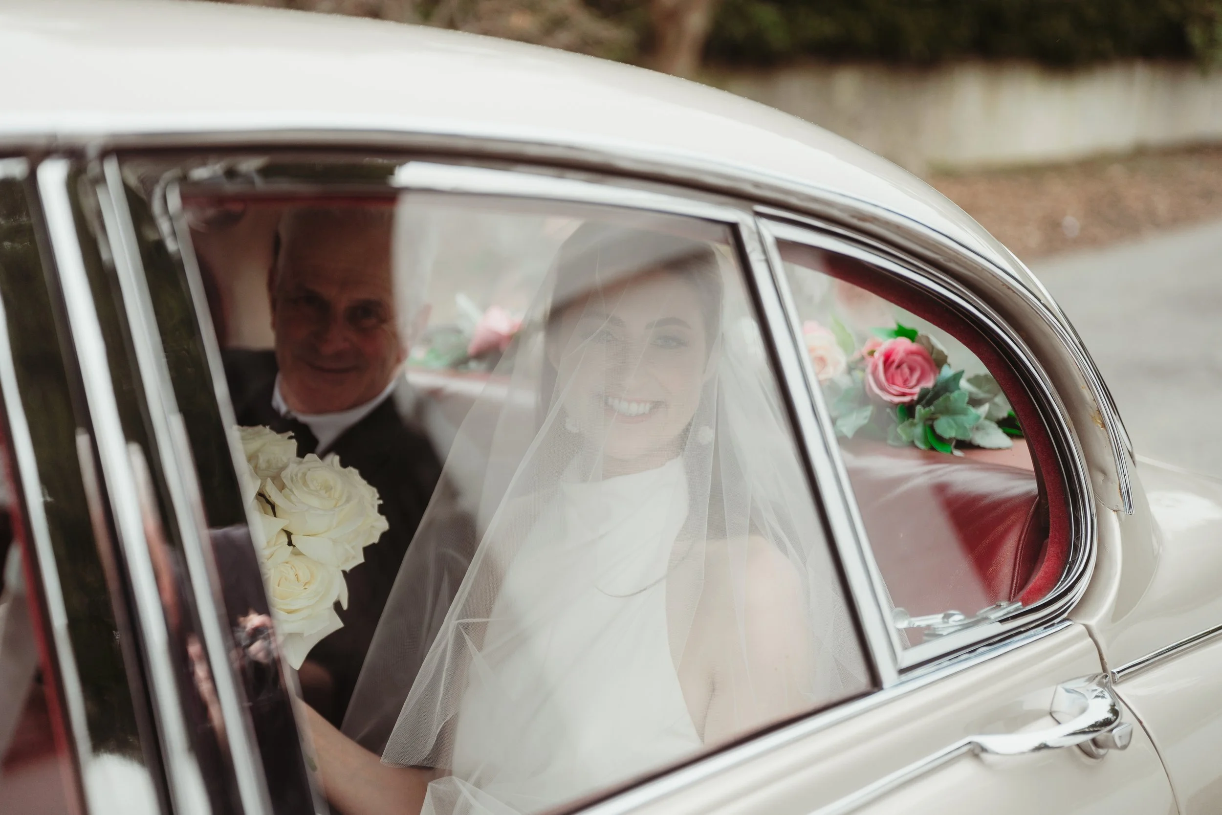 A bride in a wedding dress and veil sitting in the back of a vintage car, holding a bouquet of white roses, with a smiling man in a suit sitting beside her. The car interior features red upholstery and flowers.