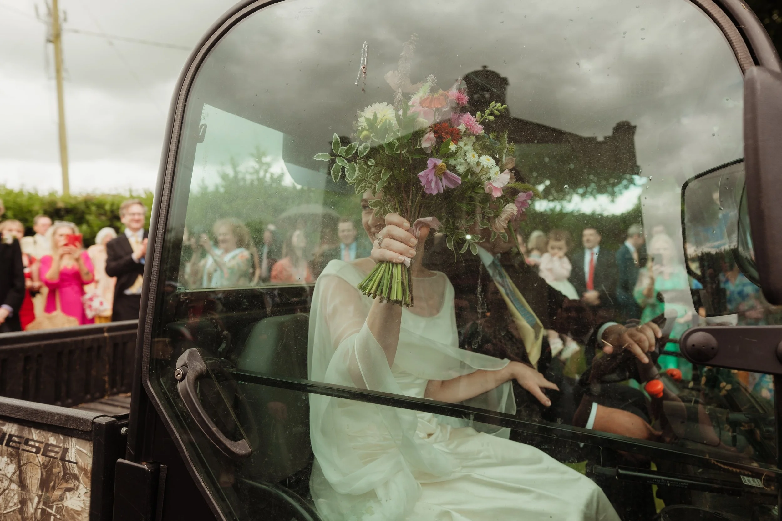 A bride holding a bouquet of flowers inside a vehicle during a wedding celebration, with guests in formal attire visible outside in the background.