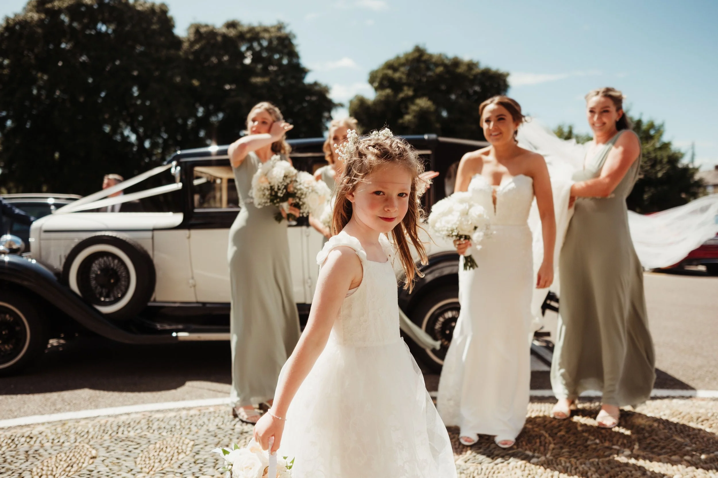 A young girl in a white dress standing in front of a vintage black and white car, with four women in wedding dresses and silver dresses behind her, holding bouquets of white flowers.