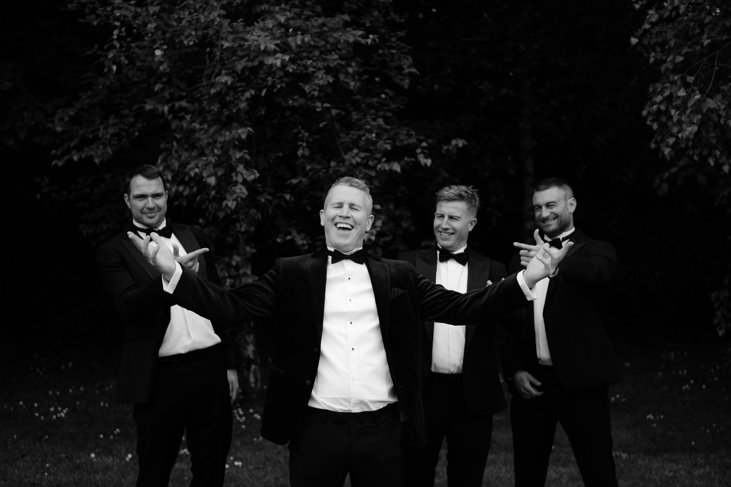 Four men dressed in tuxedos and bow ties, smiling and posing outdoors at night, with trees in the background.