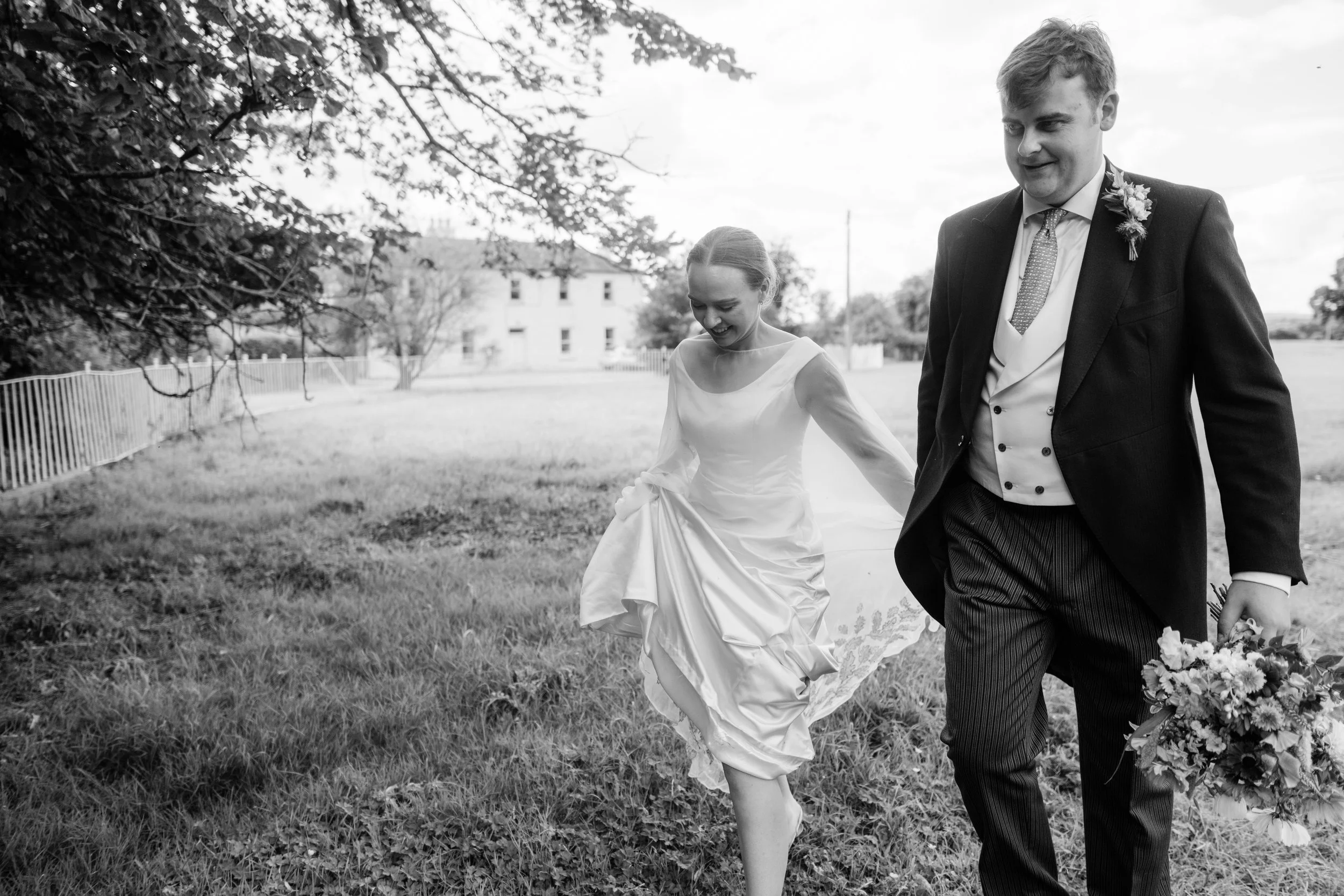 Black and white photo of a bride and groom walking outdoors on grass, smiling and holding hands, with the bride lifting her dress slightly.