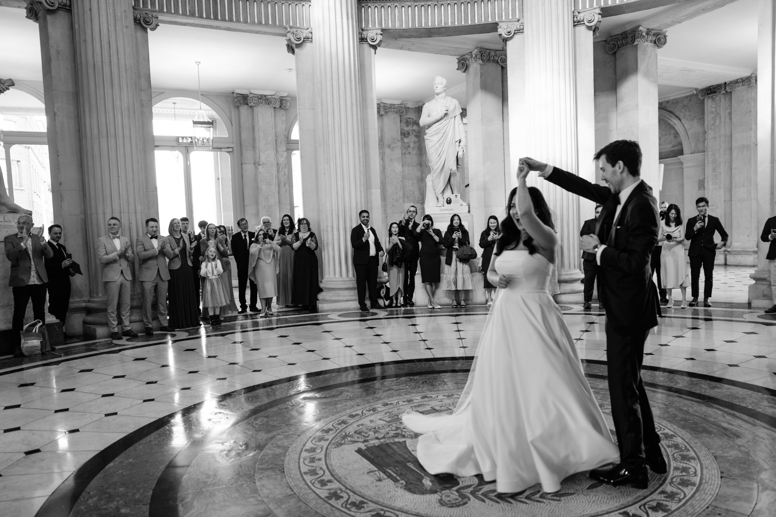 A bride and groom dance in the center of a grand hall with classical columns and a statue, while an audience watches and takes photos.