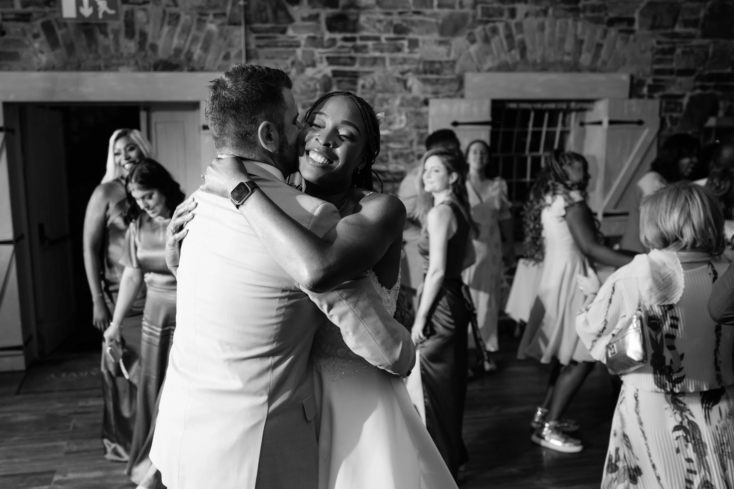 A couple dancing closely at a wedding reception, the man in a light-colored suit and the woman in a wedding dress, smiling happily, with other guests dancing and socializing in the background.