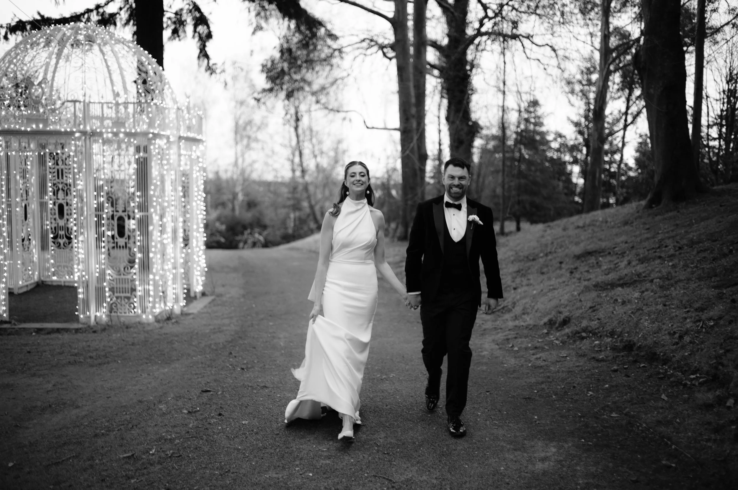 Black and white photo of a smiling bride in a wedding gown and a groom in a tuxedo holding hands and walking outdoors near a lit decorative gazebo surrounded by trees.