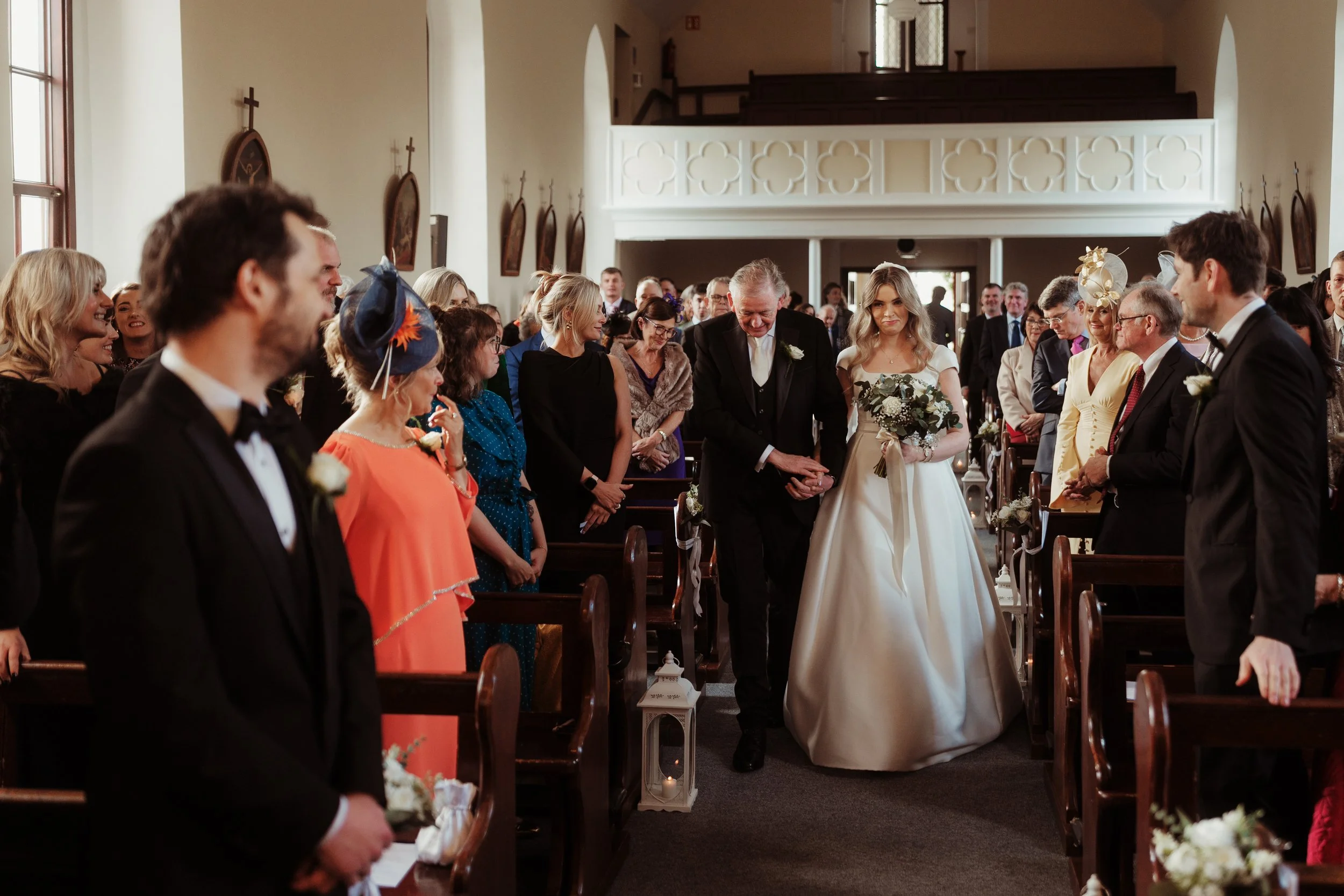 Bride walking down the aisle with her father during a wedding ceremony in a church, surrounded by seated guests on both sides.