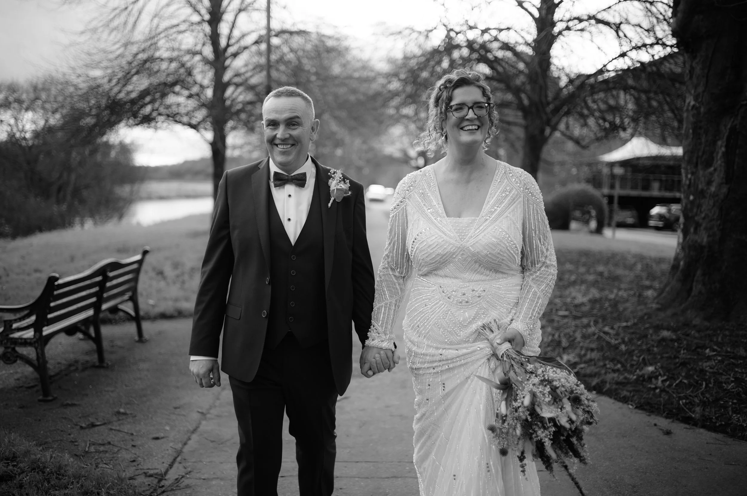 A black and white photo of a happy bride and groom walking hand in hand outdoors, with trees and park benches in the background.