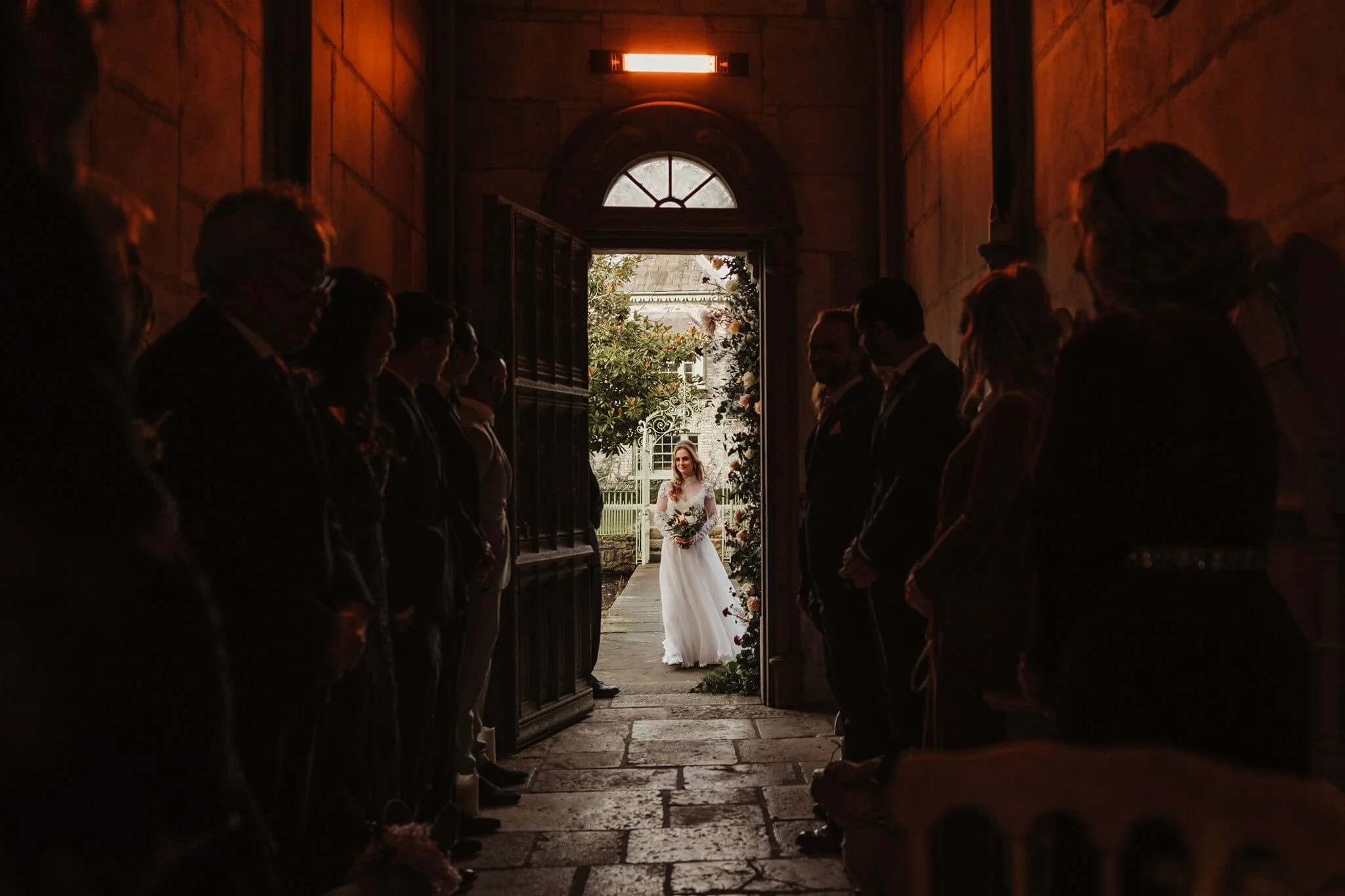 Bride standing at an open doorway inside a church, holding a bouquet, as guests look on from darkened interior on each side.