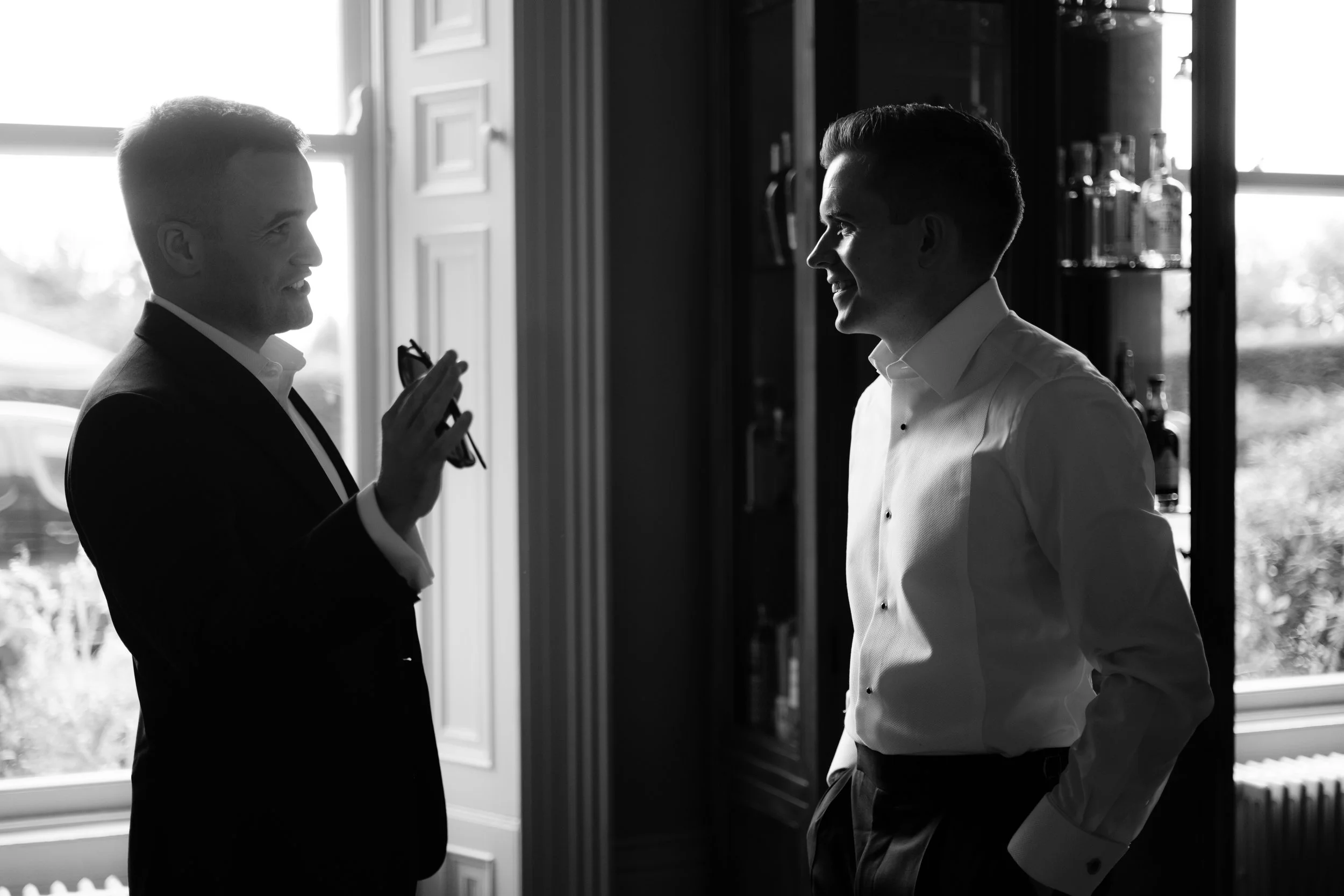 Two men in formal attire, smiling and talking in a room with large windows, black and white photo.