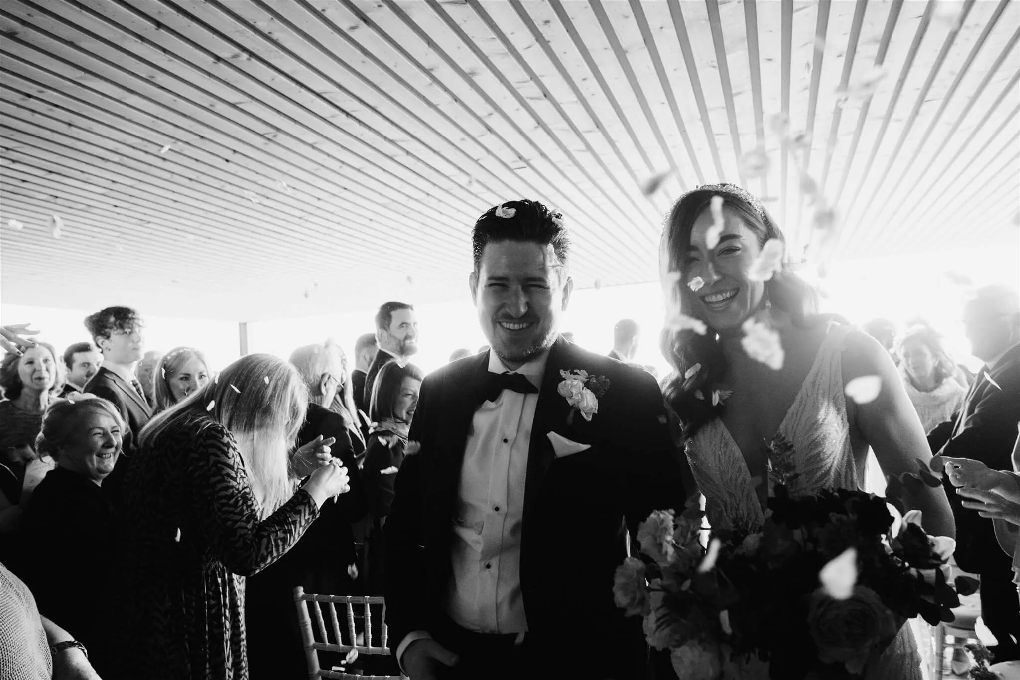 A black-and-white photo of a wedding celebration, with a bride and groom smiling and confetti falling around them. The bride holds a bouquet of flowers, and the groom wears a tuxedo. Guests are in the background, celebrating and cheering.