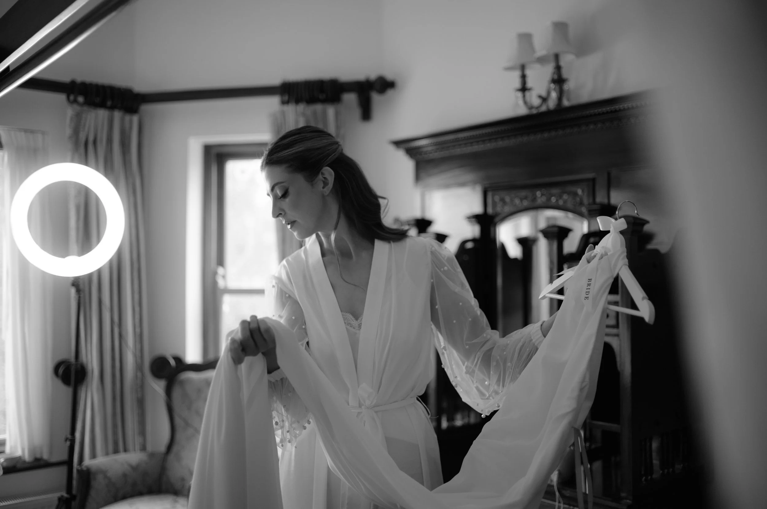 A bride in a wedding gown looking at a dress on a hanger in a bedroom with vintage decor.