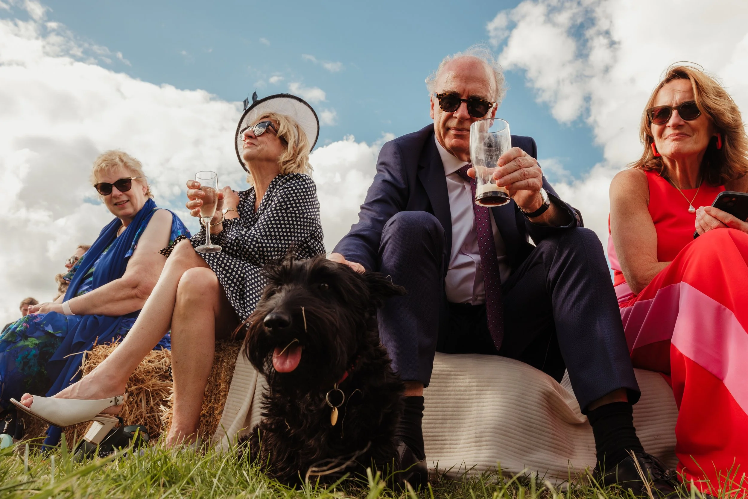 Group of elderly people sitting outdoors on hay bales at a sunny event, with a black dog in the foreground, some holding drinks, dressed in colorful and formal attire, against a backdrop of a partly cloudy sky.