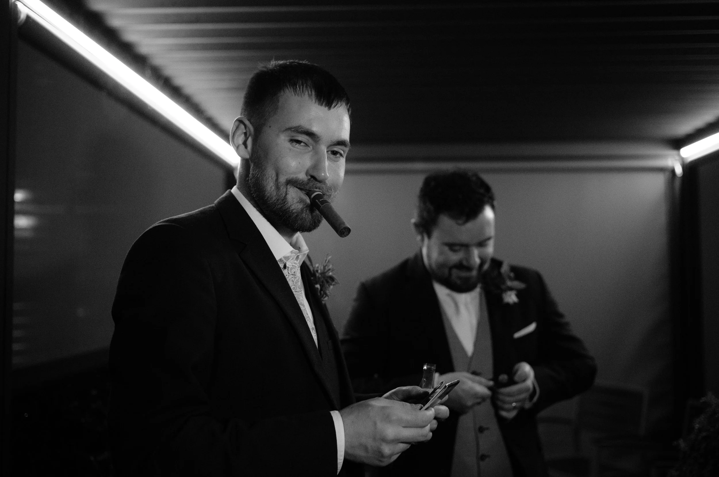 Two men in suits, one with a cigar, smiling while looking at their phones at a formal event.