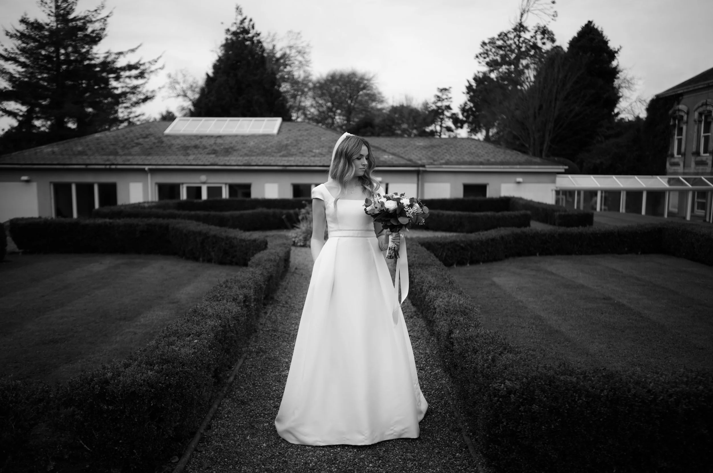 Black and white photo of a woman in a wedding dress holding a bouquet, standing on a garden pathway with neatly trimmed hedges, trees, and a building with a roof and solar panels in the background.