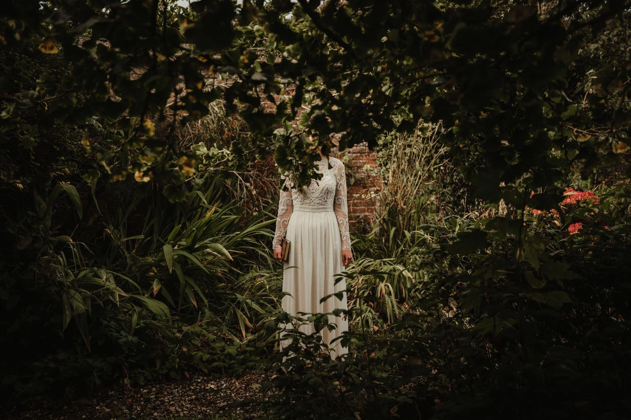 A woman in a long white lace dress holding a book, standing amidst dense dark green foliage and plants in a garden, with a brick wall in the background.