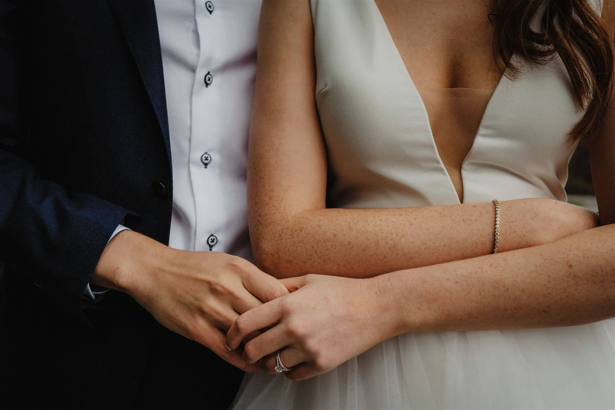 A close-up of a couple holding hands, with the woman wearing a wedding ring and a sleeveless dress, and the man in a dark suit and white dress shirt.