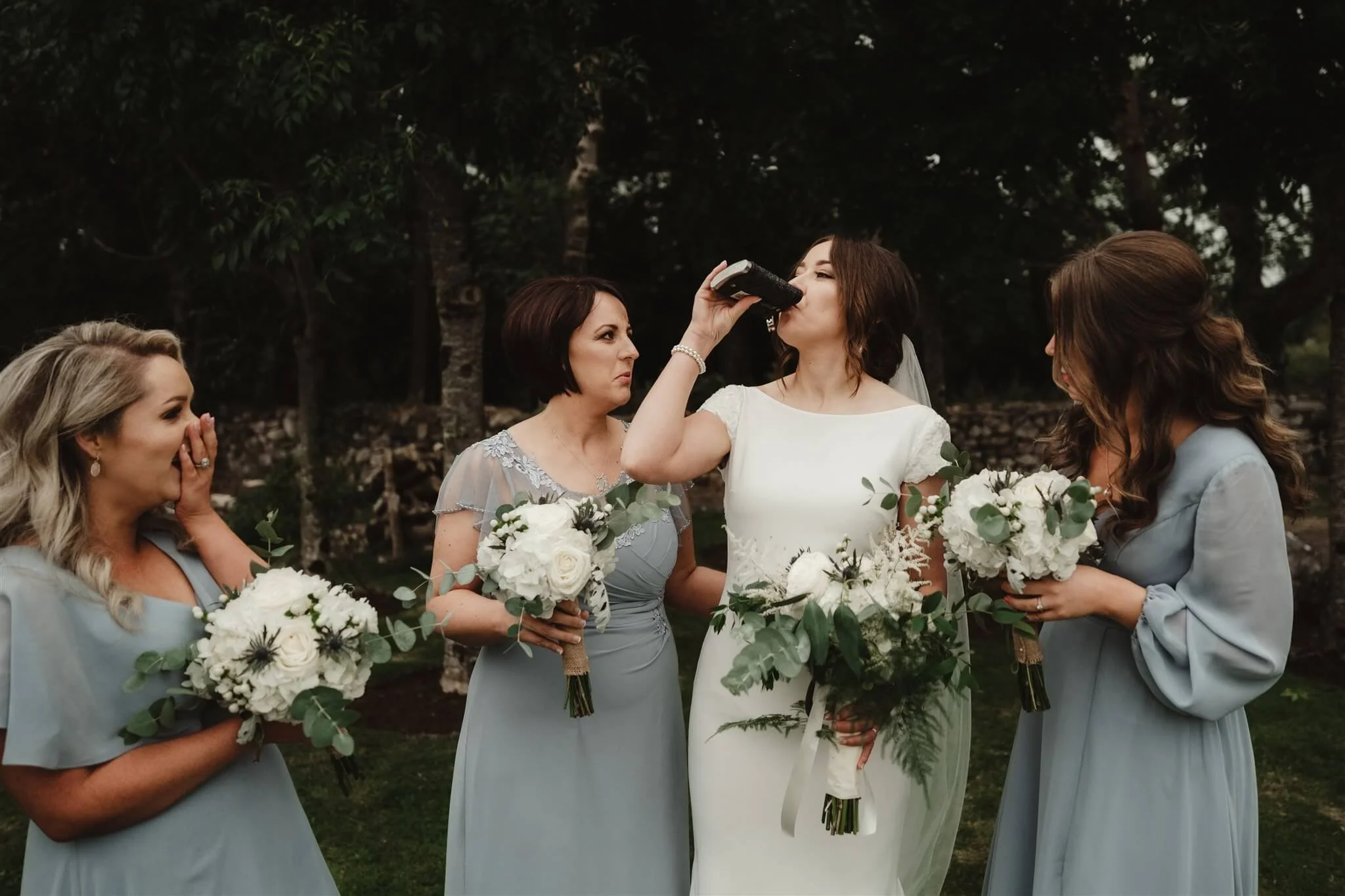 A bride and three bridesmaids standing outdoors, with the bride drinking from a water bottle while holding a bouquet. The bridesmaids are smiling and holding bouquets, wearing light blue dresses, with trees in the background.