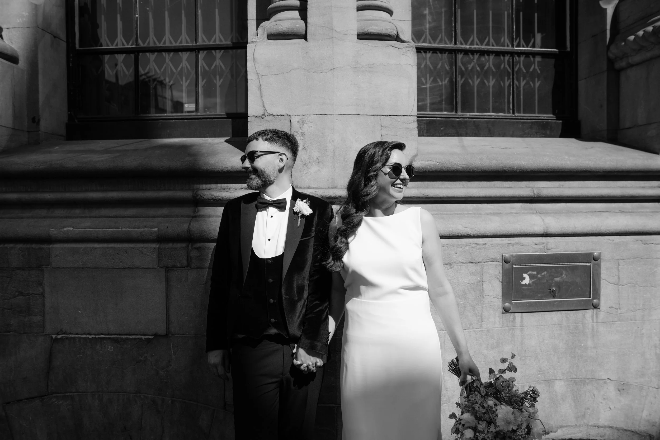 Black and white photo of a smiling bride and groom holding hands, standing against a historic building with stone steps and windows, dressed in wedding attire.
