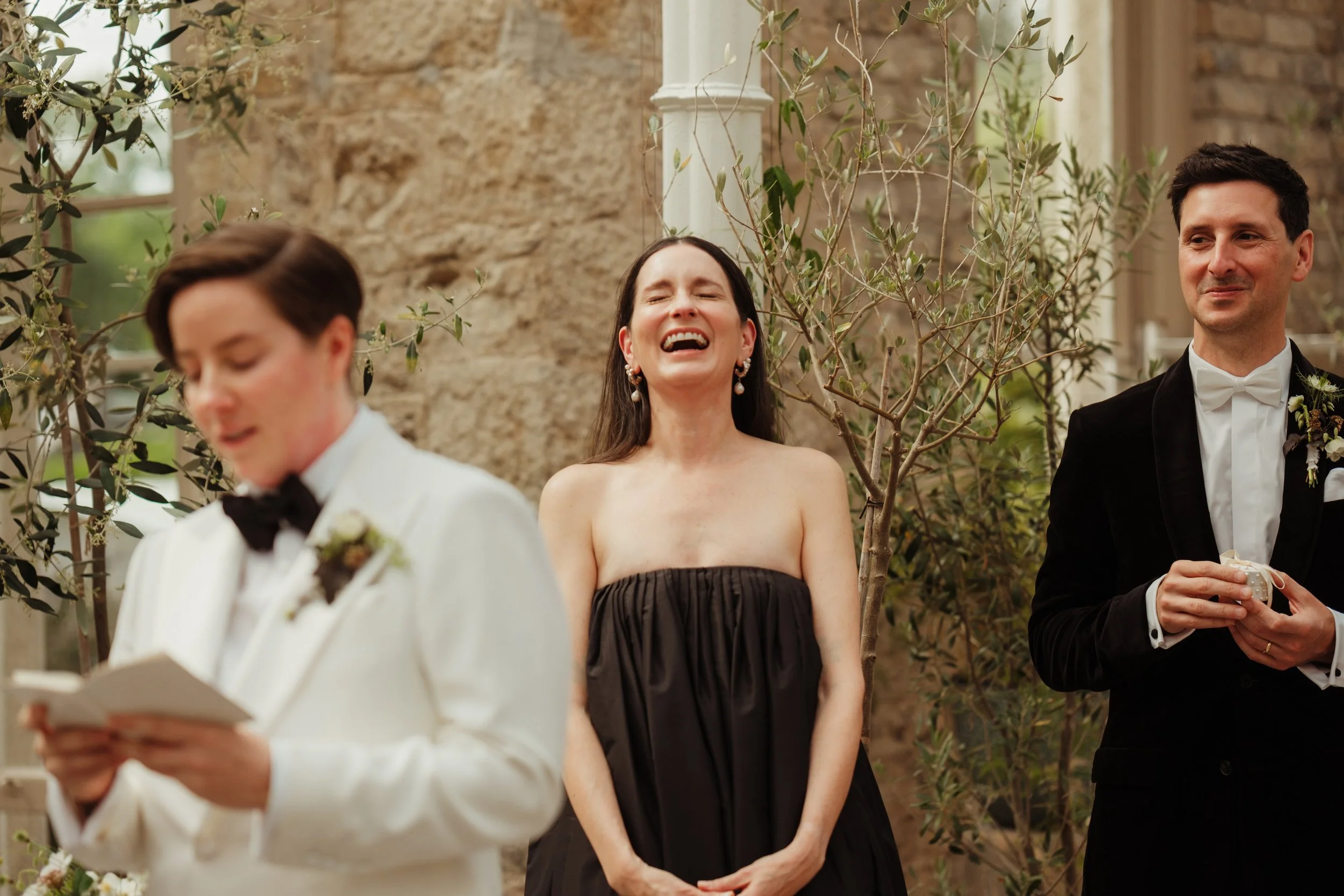 A woman in a black strapless dress is laughing with her eyes closed, standing between two men dressed in tuxedos at a wedding ceremony outdoors, with a stone wall and plants in the background.