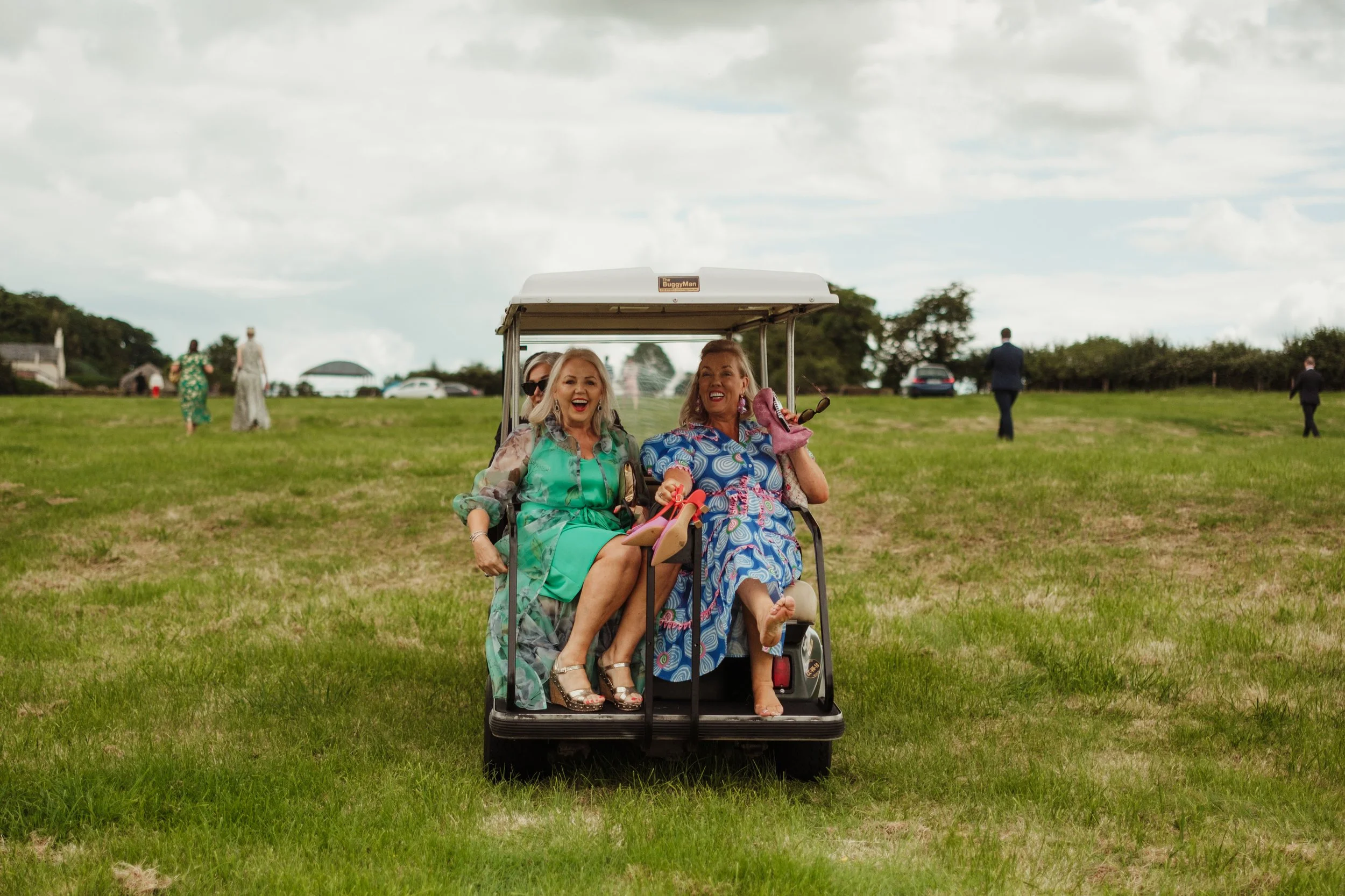 Two women in colorful dresses sitting in a golf cart on a grassy field, smiling and enjoying themselves.