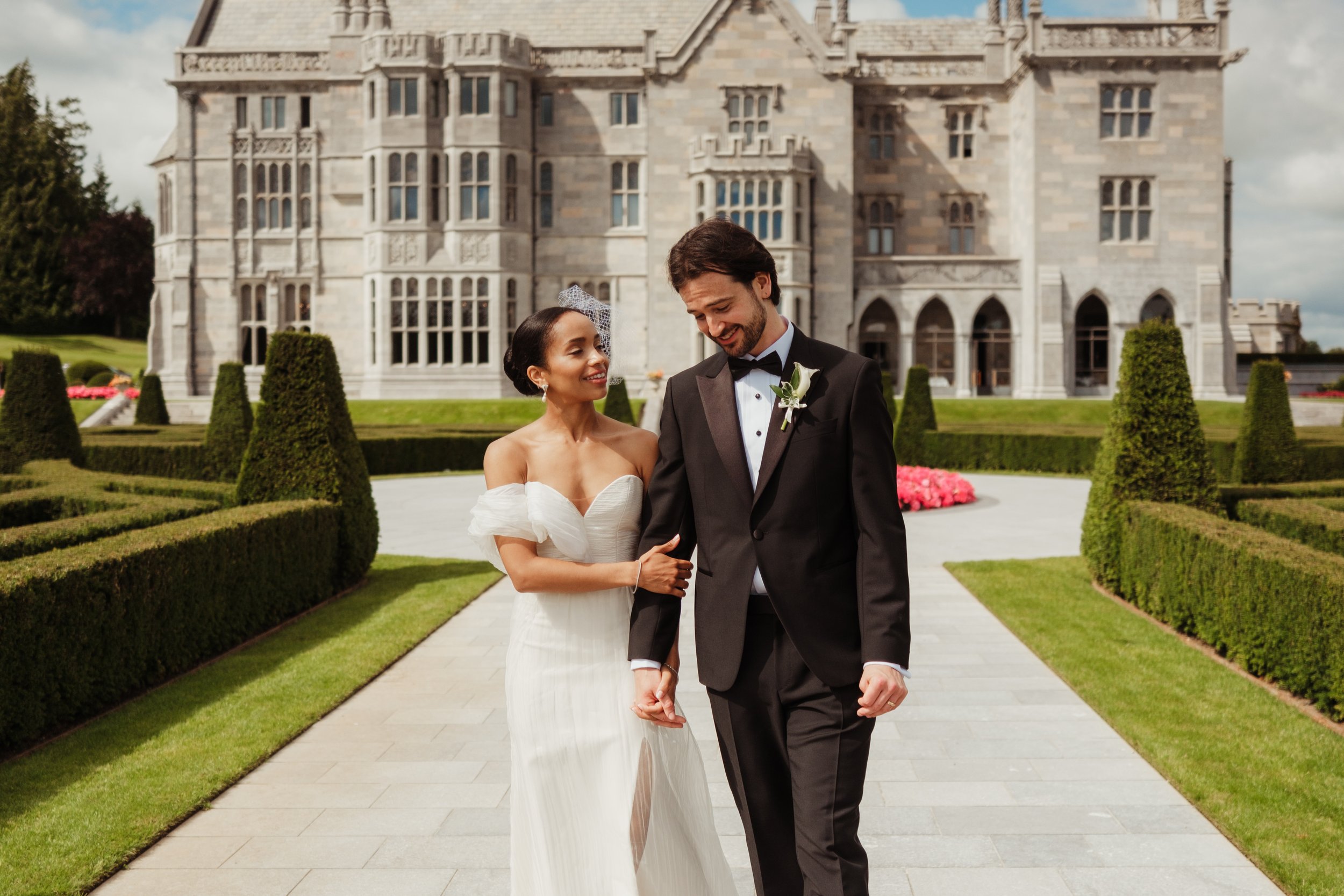 A newlywed couple walking hand in hand outside a grand castle-like building with manicured gardens and hedges.