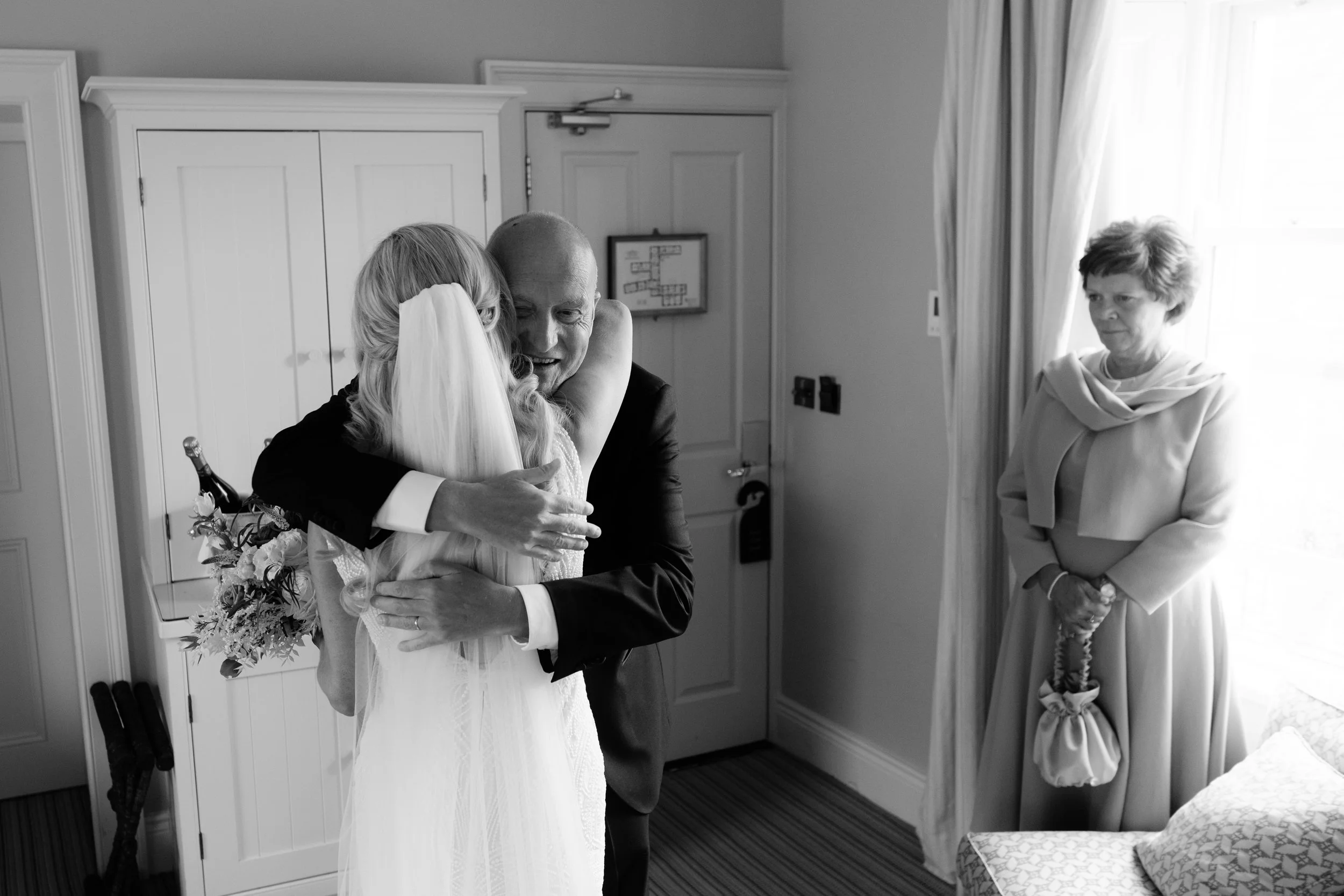 A bride hugging an elderly man, presumably her father, during a wedding moment. A woman stands nearby by a window watching them, holding a purse.