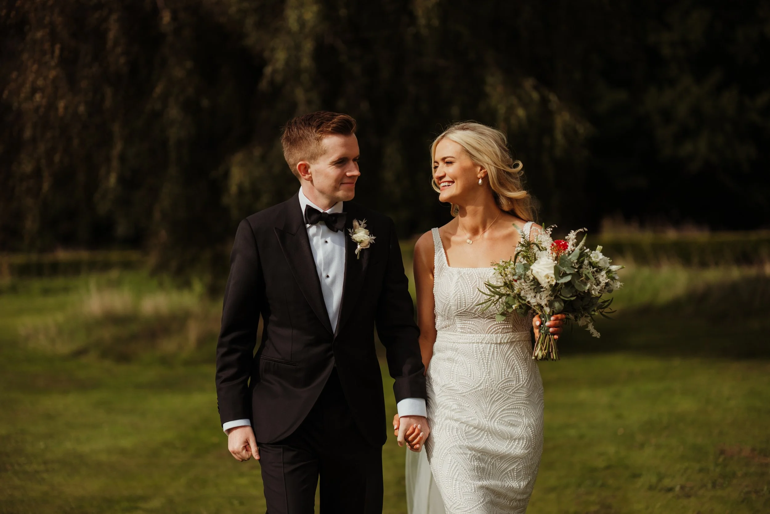 A newlywed couple walking hand-in-hand outdoors, the groom in a black tuxedo and the bride in a white lace wedding dress holding a bouquet of white and red flowers, smiling at each other.