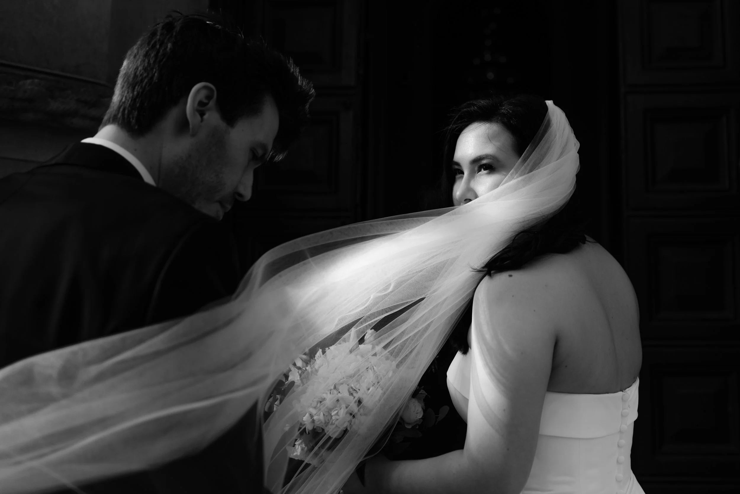Black and white photo of a bride with a veil covering part of her face, looking at a groom with his head bowed, in an intimate moment.