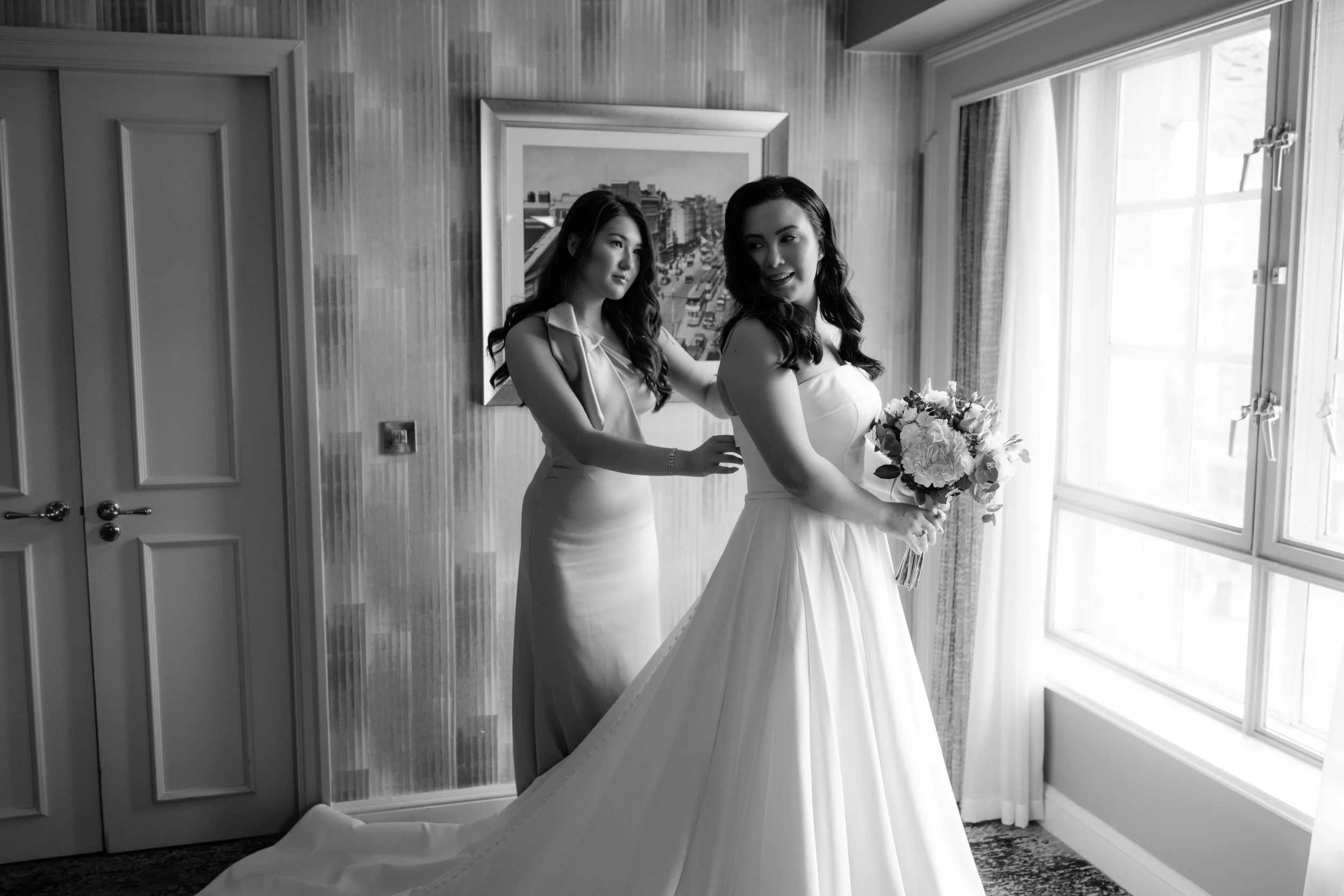A bride in a wedding dress holding a bouquet stands near a window, with a woman in a dress helping her in a room with artwork on the wall and a patterned carpet.
