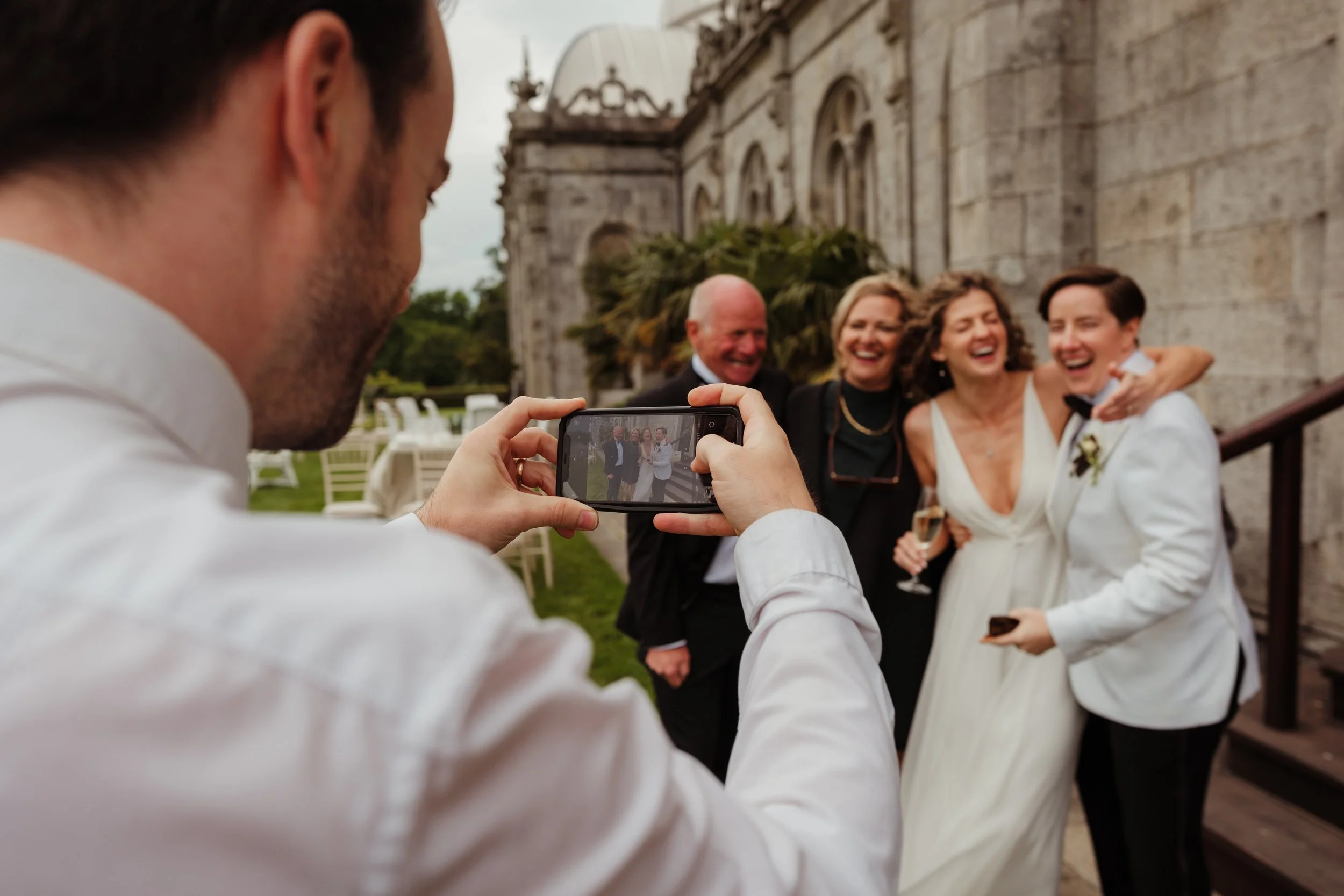 Man taking a photo of four people, including a bride and groom, outside a historic building with arches and stone walls, celebrating with smiles and drinks.
