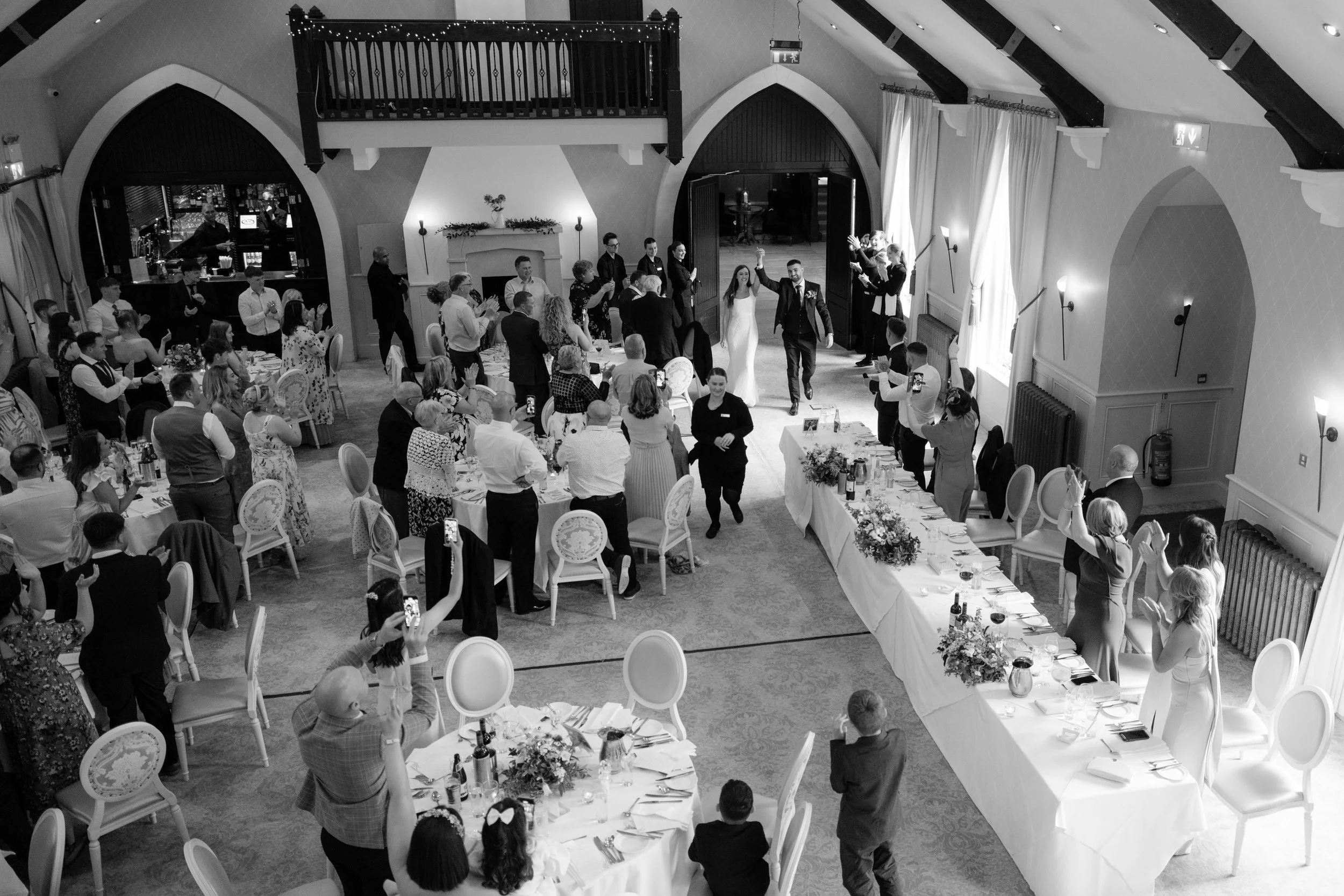 A black-and-white photo of a wedding reception in a large decorated hall with guests standing around tables, watching the bride and groom enter the room.