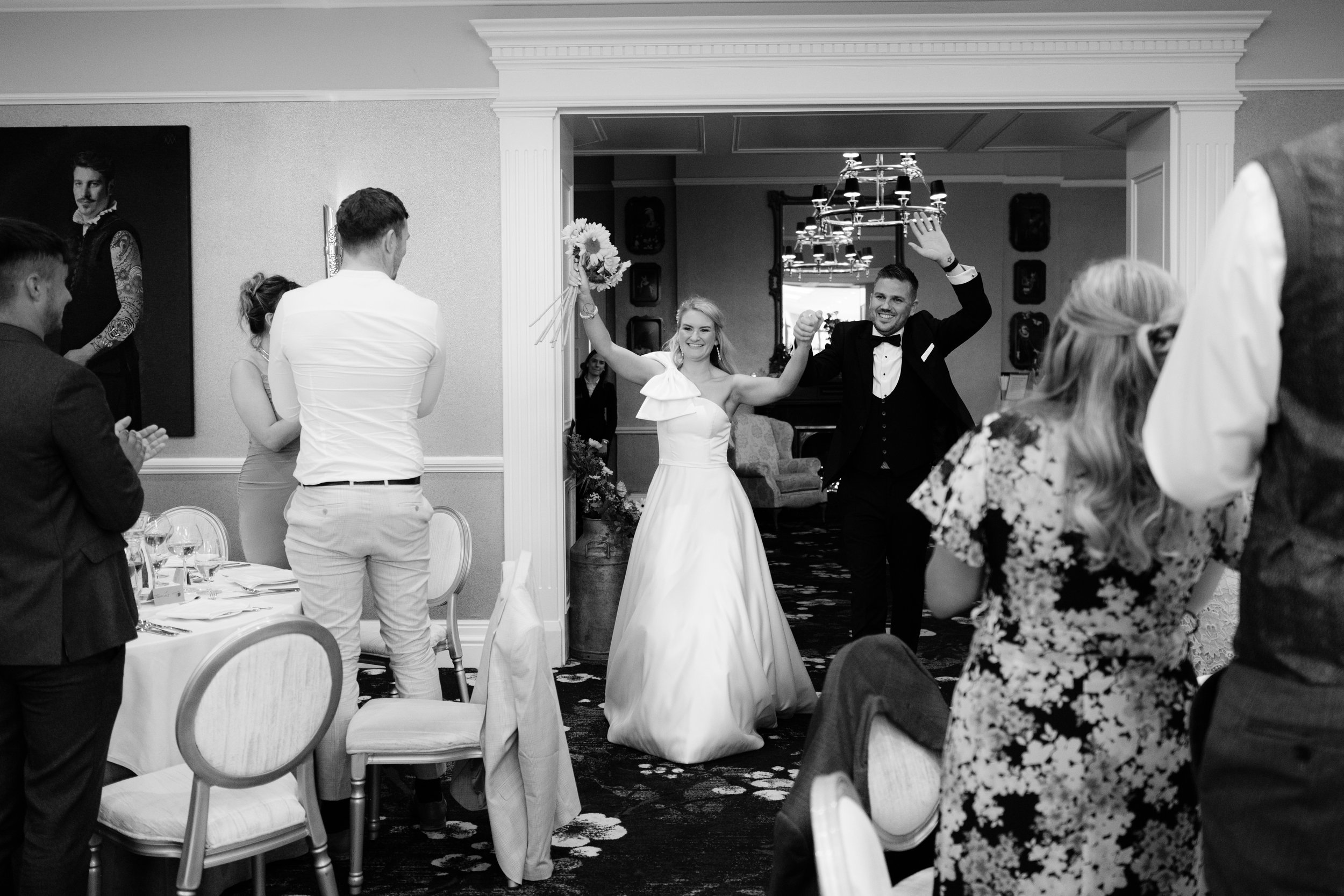 Bride and groom entering a reception hall, smiling and celebrating, with guests clapping and taking photos