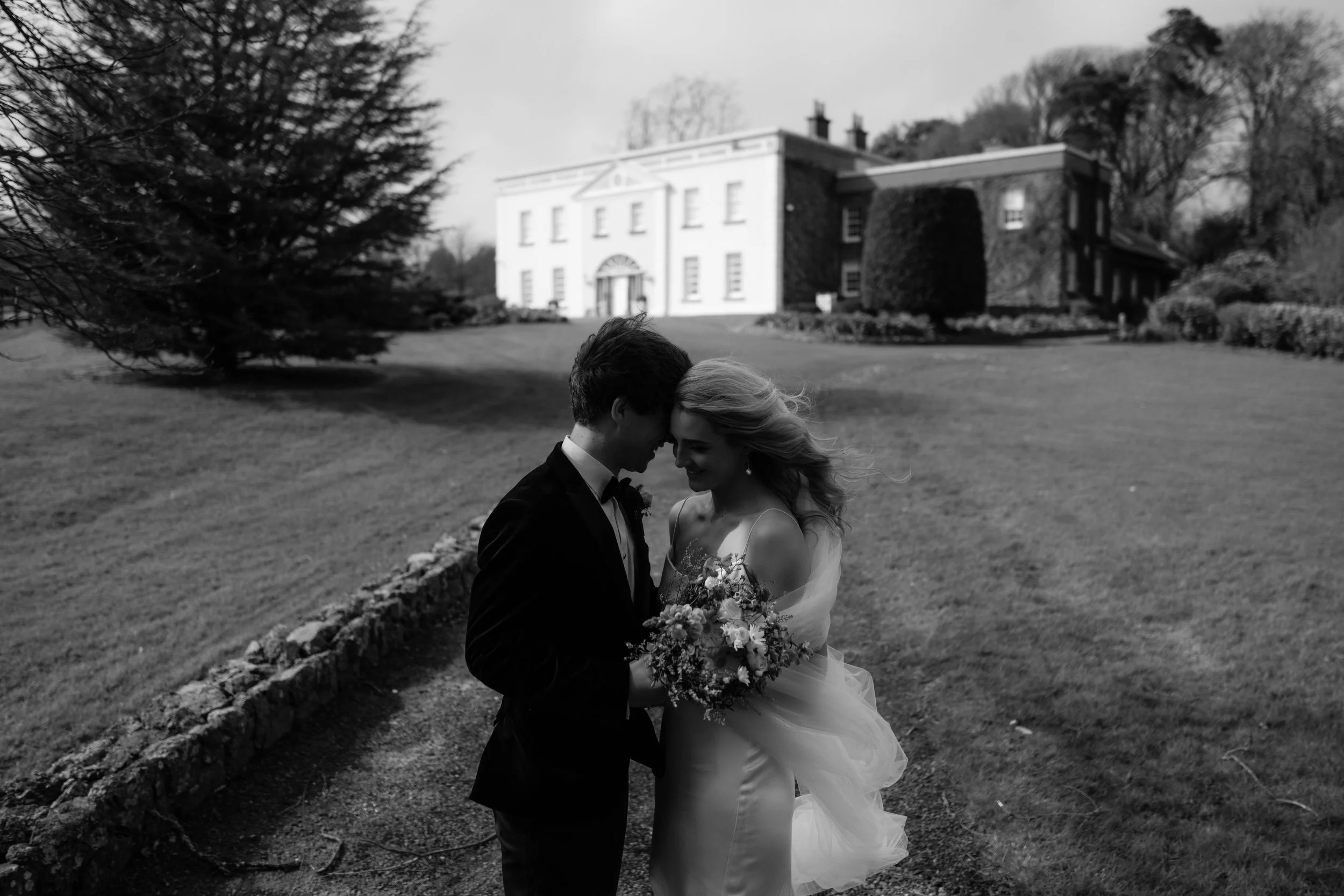 Black and white photo of a newlywed couple standing close together outdoors on a lawn, smiling and touching foreheads, with a large historic estate building in the background.