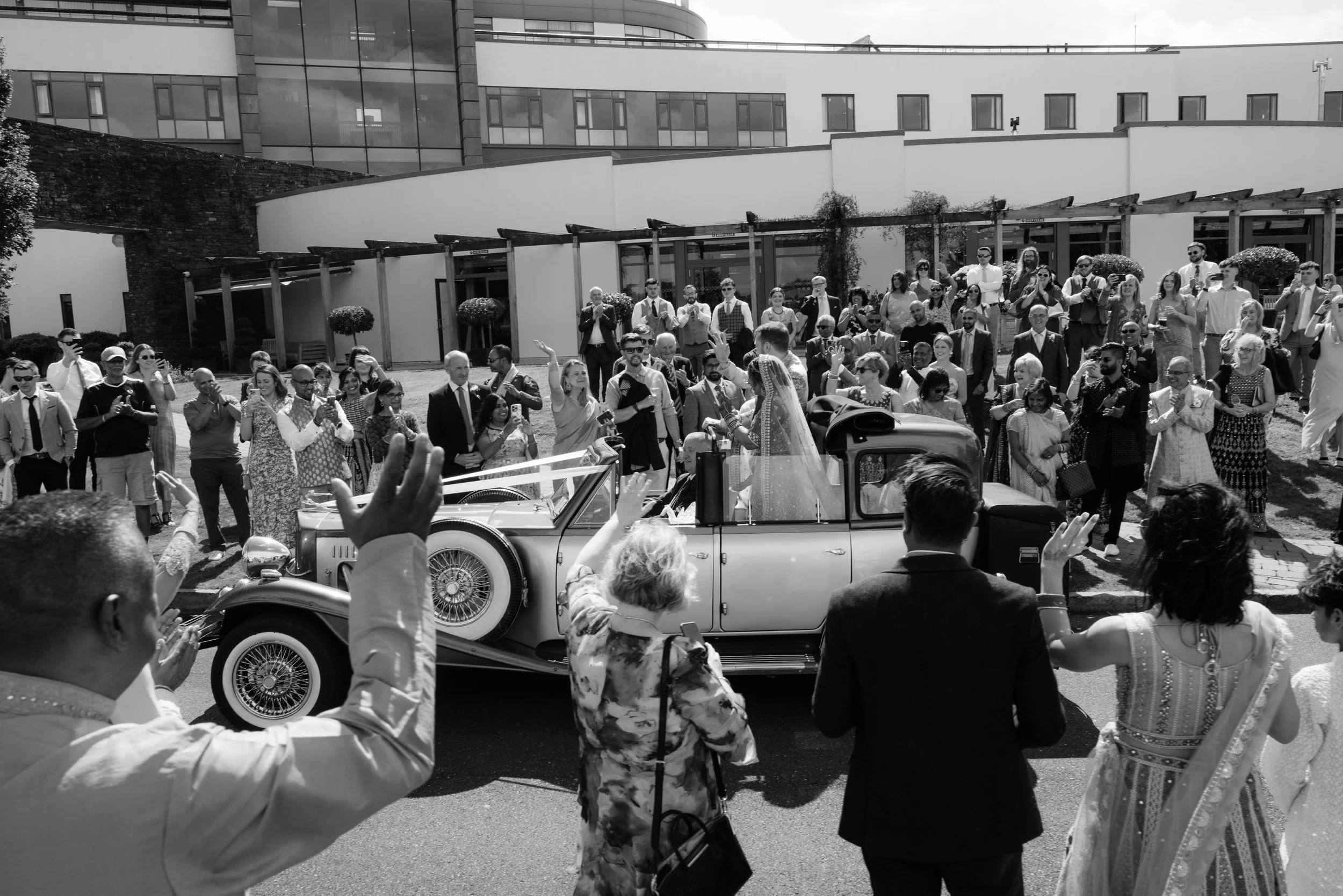 A wedding celebration with guests waving at a vintage car carrying the bride and groom. The scene is outdoors, in front of a modern building, with guests in formal attire, some taking photos and others clapping.