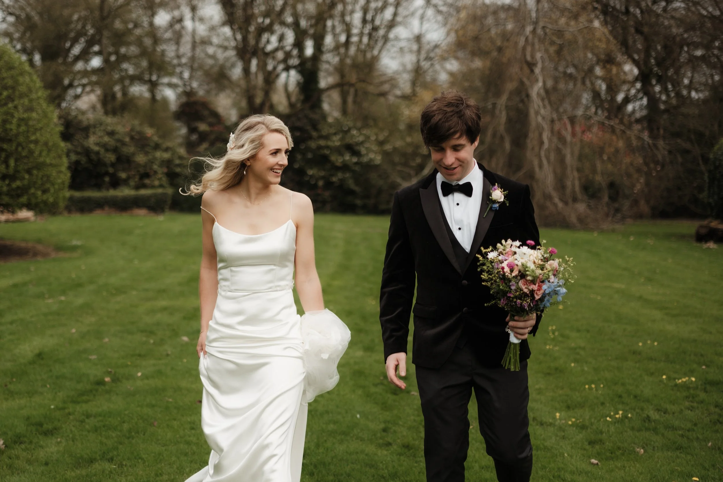 A bride and groom walking outdoors on a grassy area with trees in the background, smiling and holding hands after a wedding ceremony.