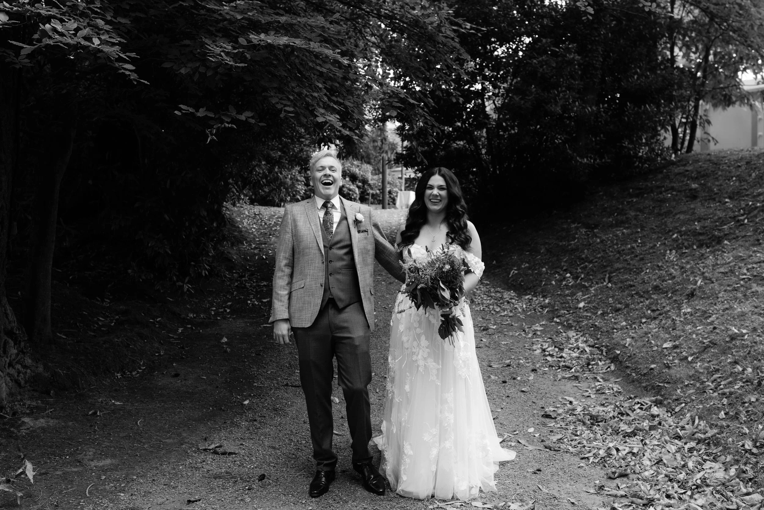 A black-and-white photograph of a bride and groom walking outdoors, happy and smiling, with the bride holding a bouquet of flowers.