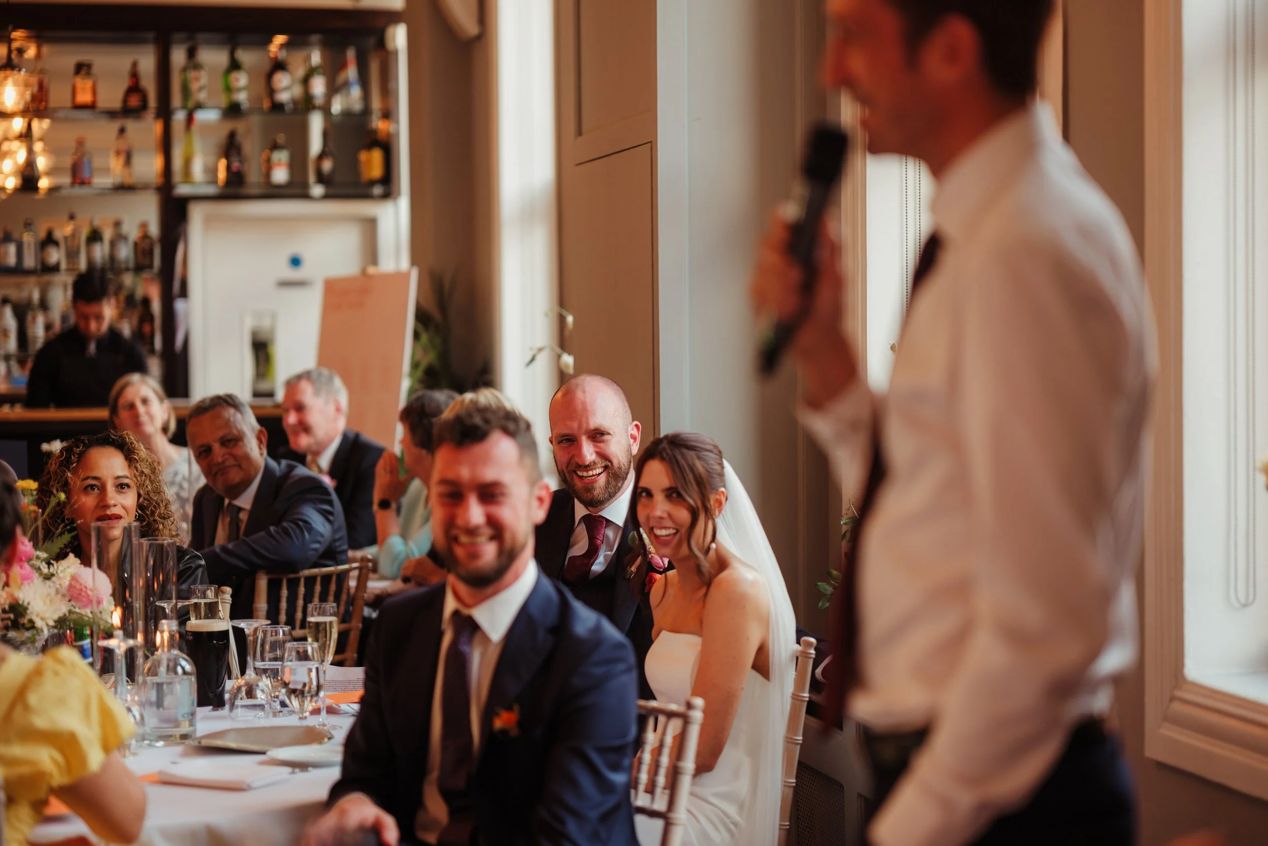 A wedding reception with guests sitting at table, listening to a man giving a speech with a microphone, including a smiling bride and groom.
