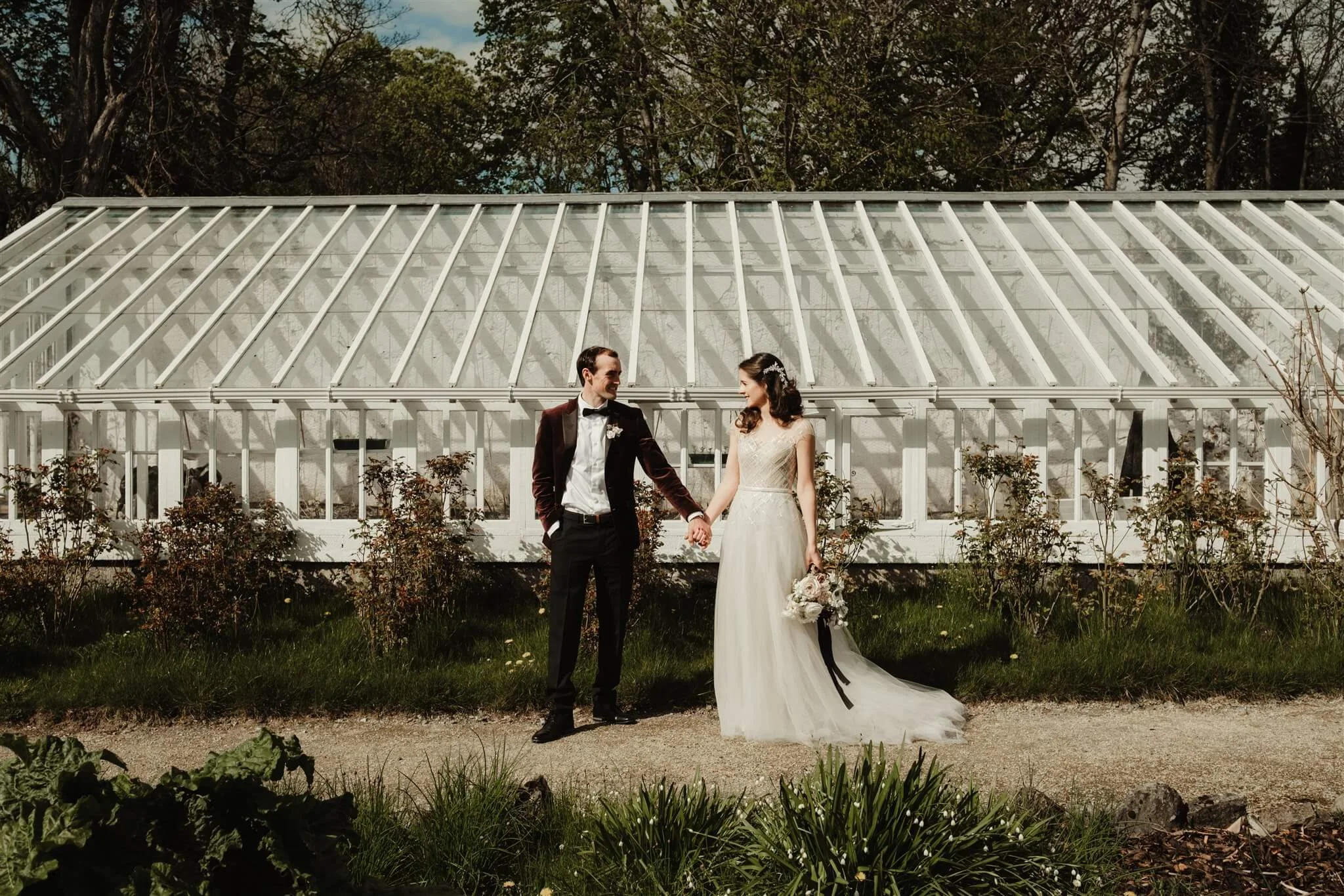 A bride and groom standing outside a greenhouse, holding hands and smiling at each other on their wedding day.