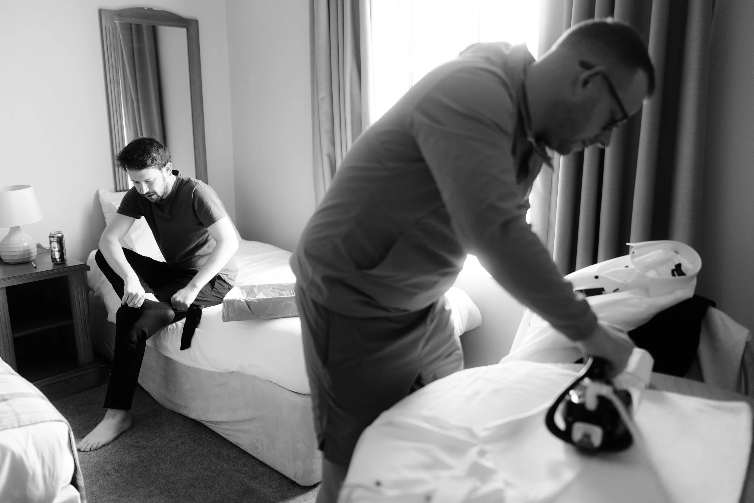 A man in glasses and a jacket is packing an iron on a bed, while a younger man in a t-shirt and pants sits on the edge of the bed, putting on socks in a hotel room. The room has a window with curtains, a lamp, and a mirror.