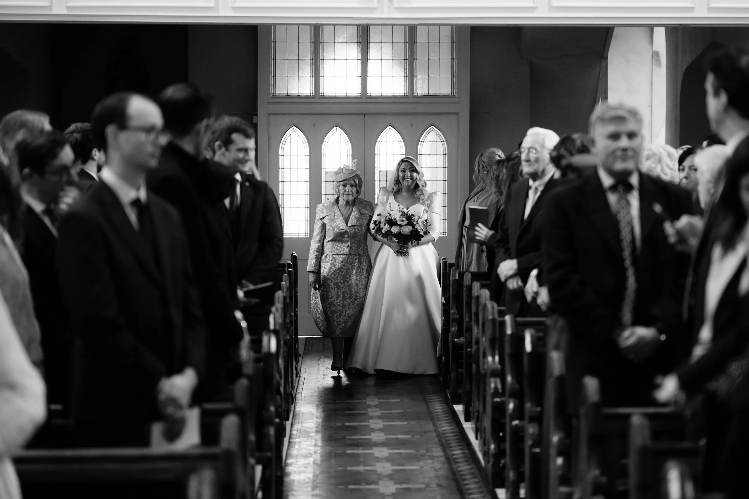 A bride walking down the aisle of a church, smiling, holding a bouquet of flowers, with an elderly woman beside her, surrounded by guests seated on both sides of the pews.