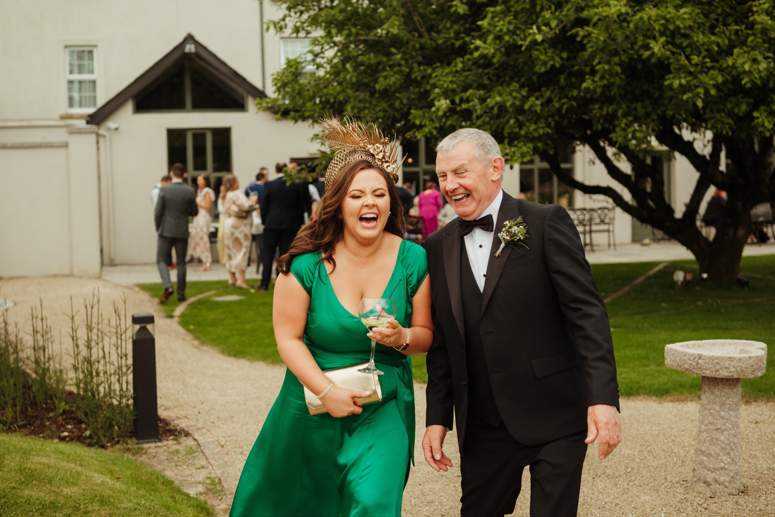 A woman in a green dress and an older man in a black tuxedo are laughing together at an outdoor social event, with other people in formal attire in the background.